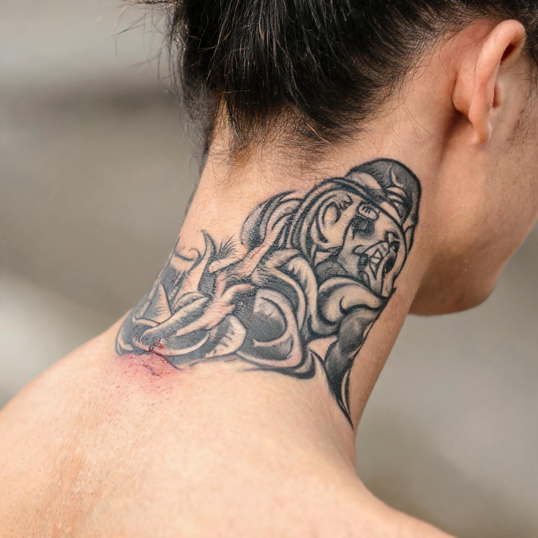 A black and grey illustrative cholo tattoo image on the nape of a neck, shown in a close-up photo against a neutral background.