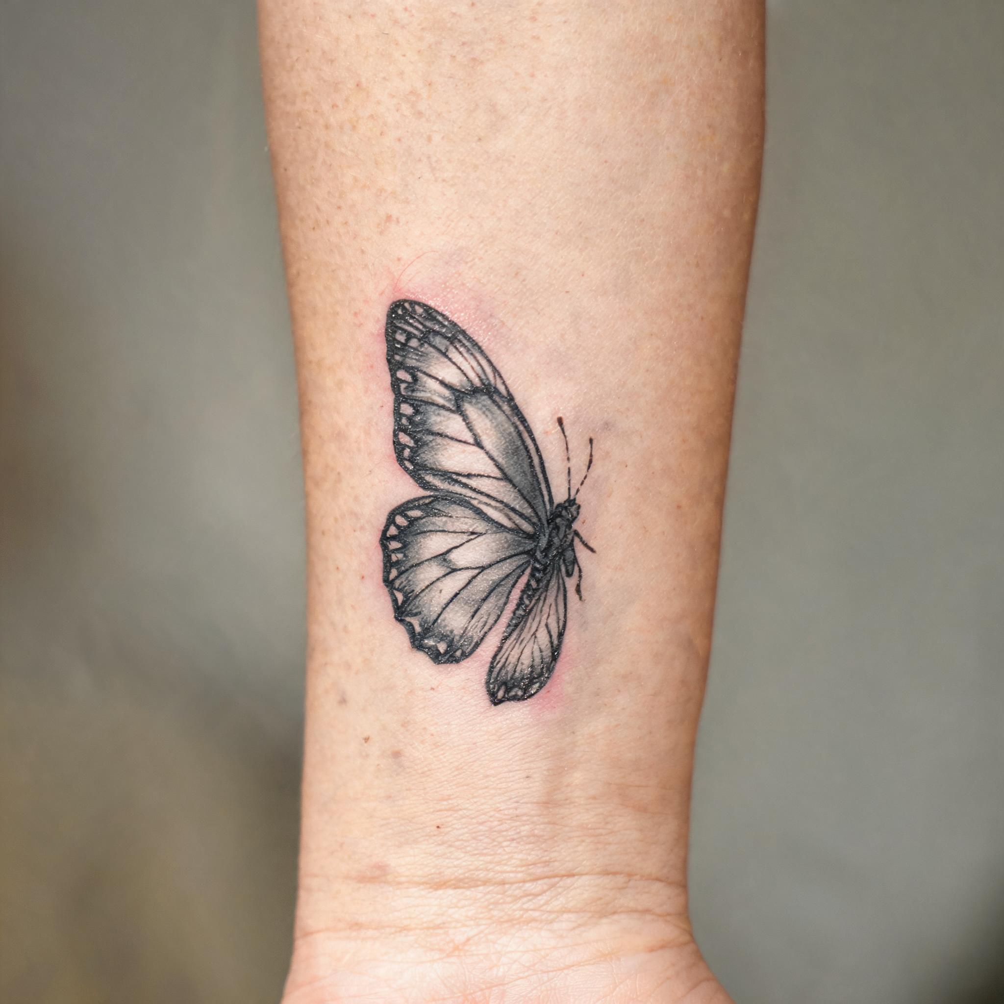 Illustrative black-and-grey butterfly tattoo images shown on an inner forearm, photographed on a clean neutral background as a tattoo image.