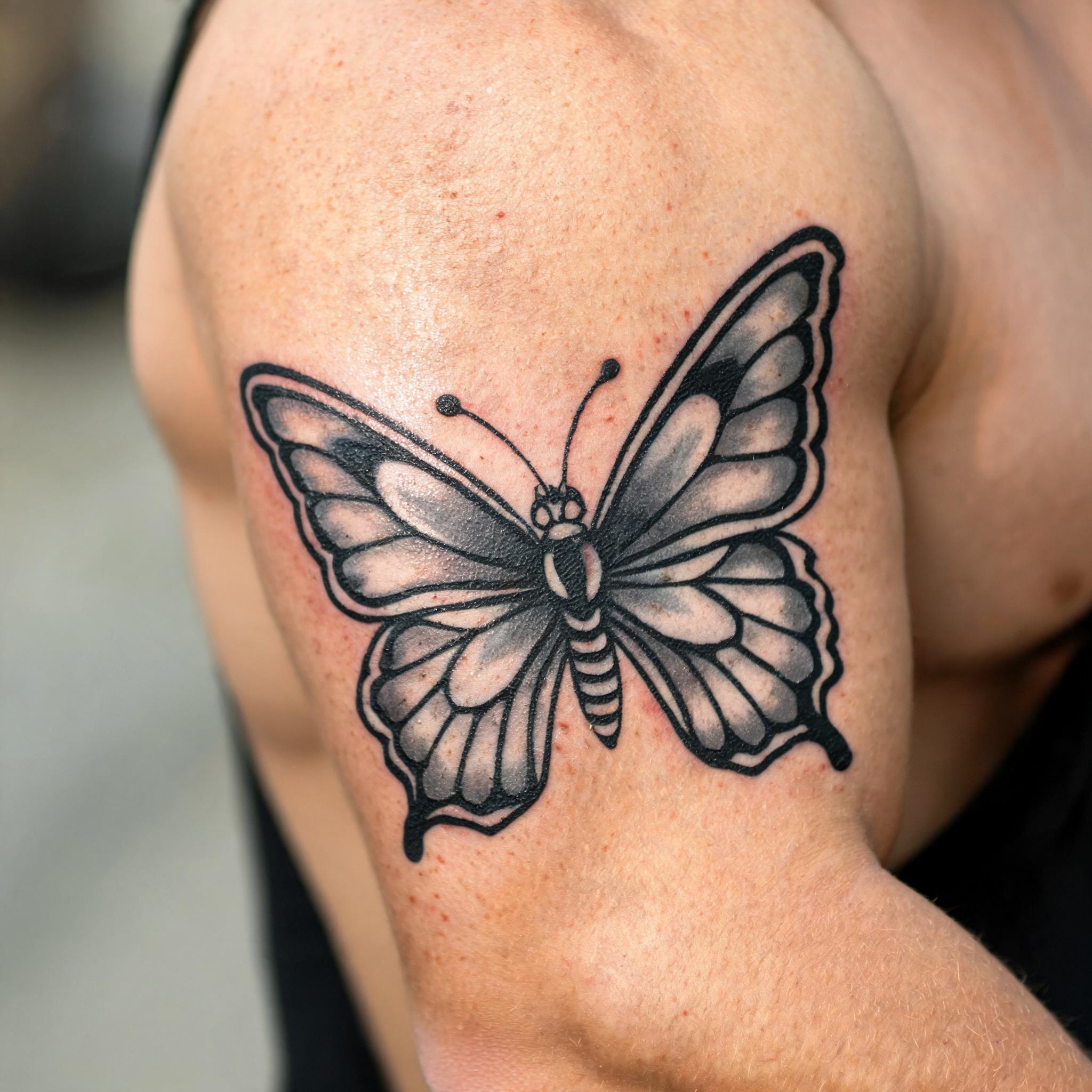 Black and grey American Traditional butterfly tattoo image on the outer upper arm, shown on clean skin against a neutral studio background.