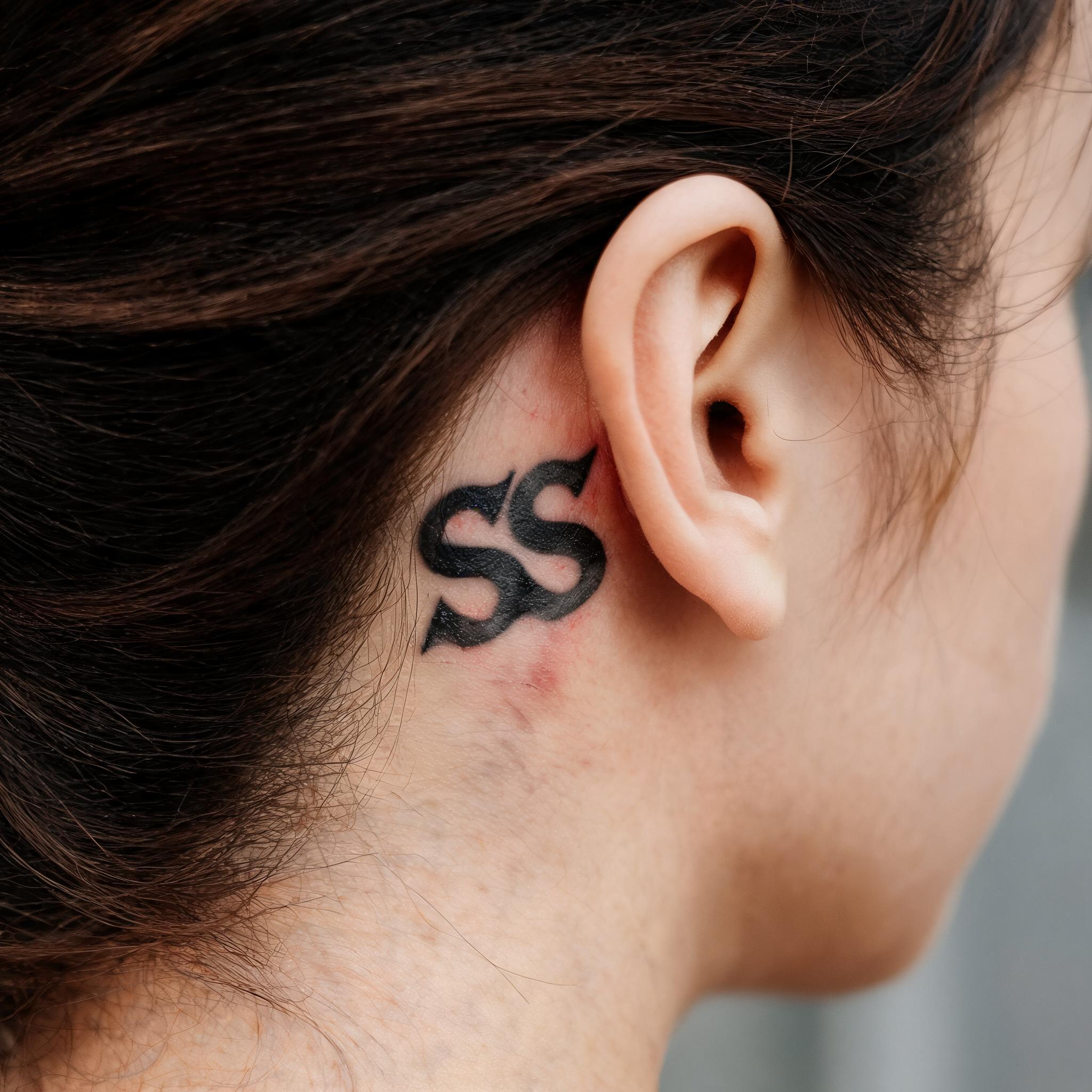 A black and grey ss tattoo images tattoo behind the ear, shown in a close-up editorial photograph on natural skin.