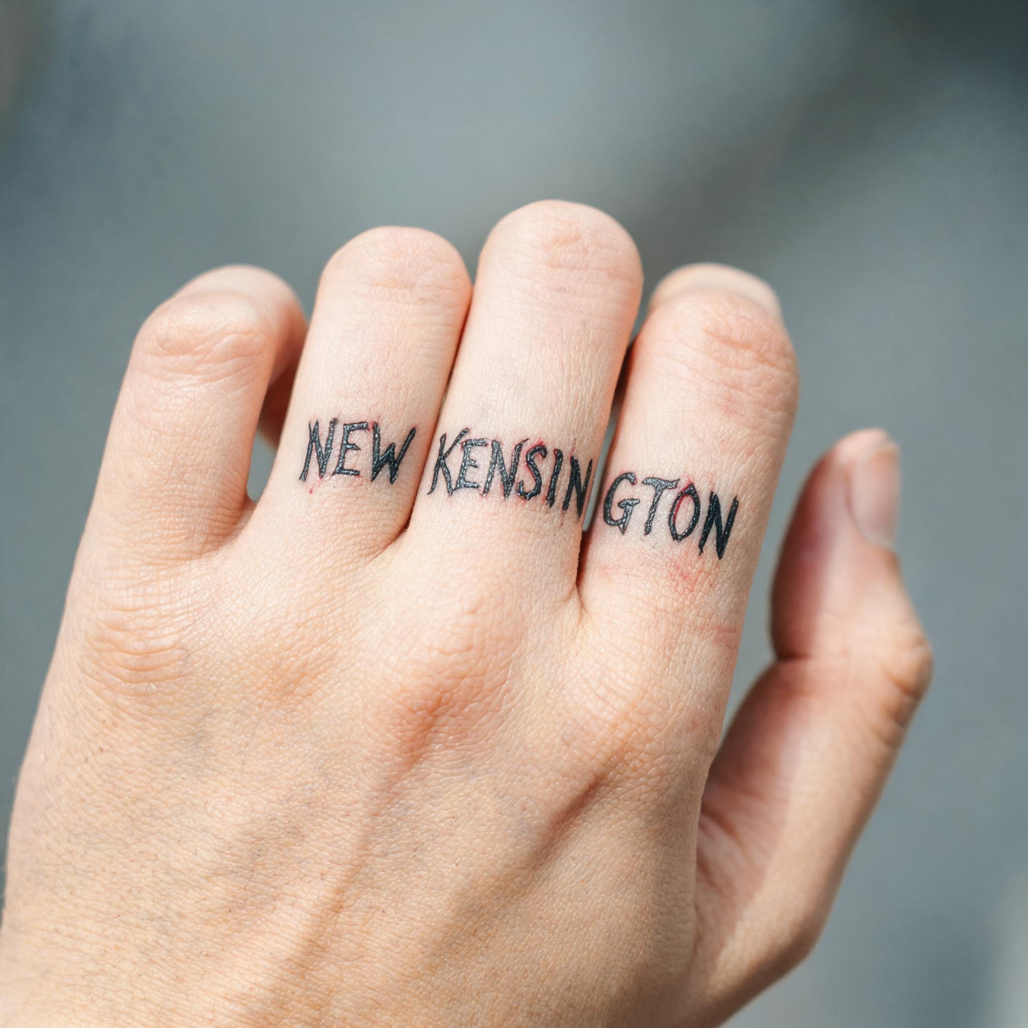 Black and grey illustrative tattoo of a true image design on a finger, shown on a relaxed hand against a neutral background.