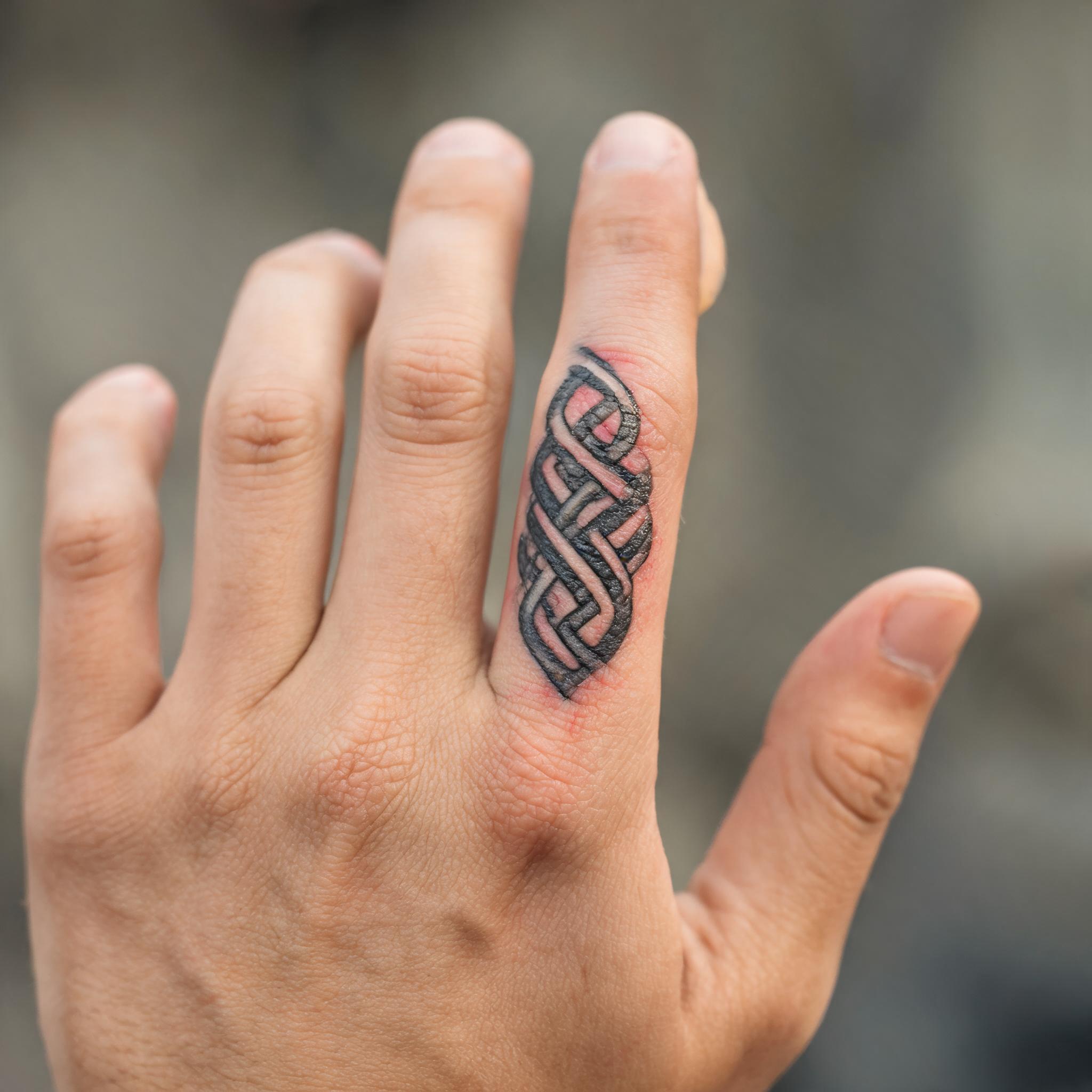 A close-up tattoo image of a Celtic knot inked along a person's finger, shown in black and grey realism on a white background.