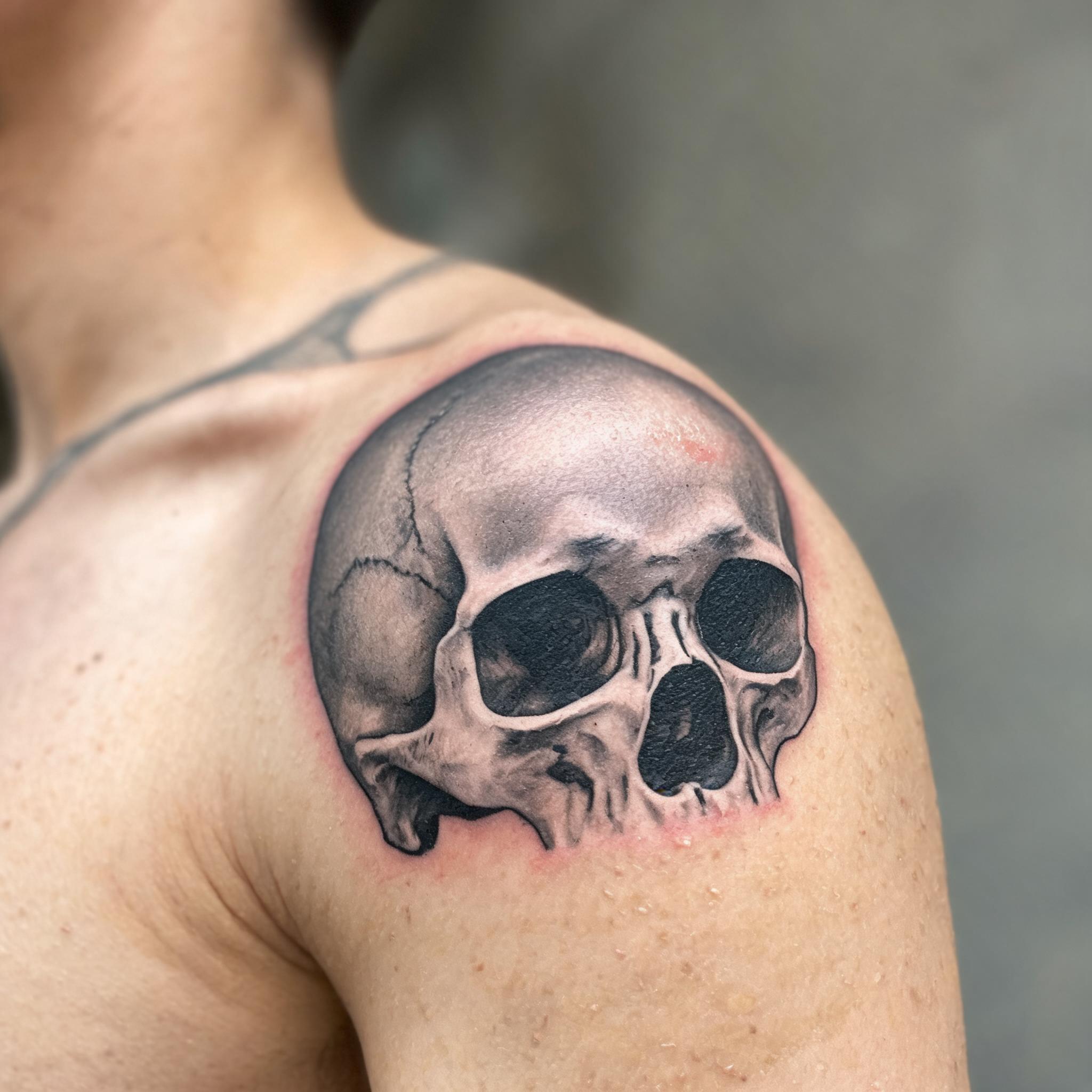 A black and grey realism tattoo image of a skull on a shoulder cap, shown on skin against a clean neutral background.