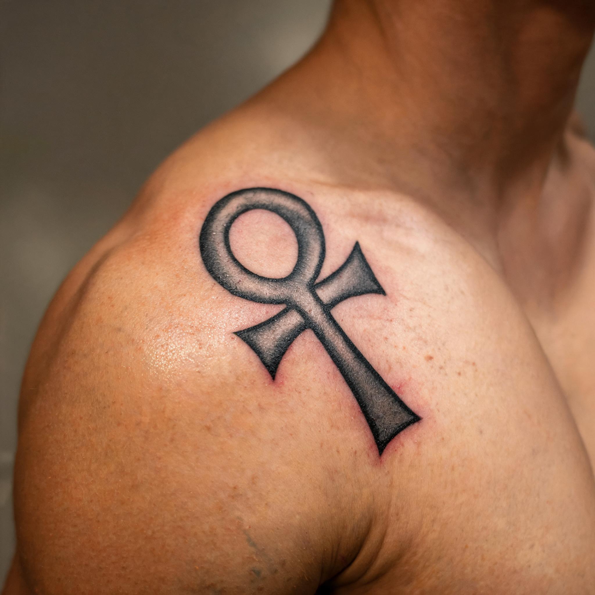 A black and grey realism tattoo of an ankh on a shoulder cap, shown as a tattoo image on white background.