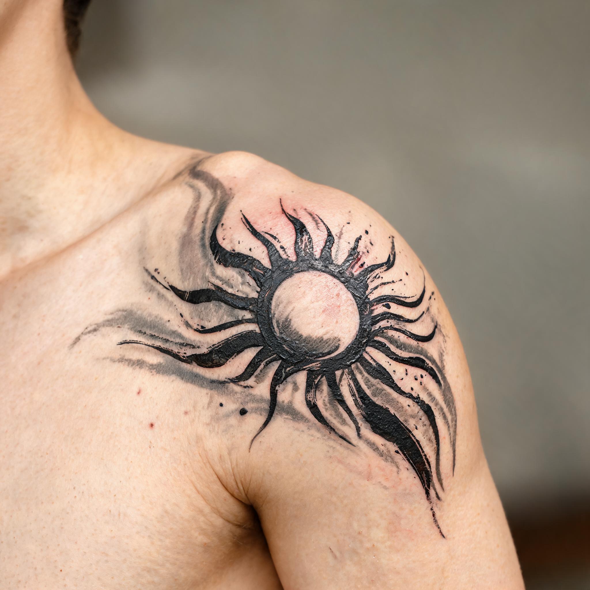A black and grey tribal sun tattoo image on a shoulder, shown in trash polka style with bold strokes and splatter.