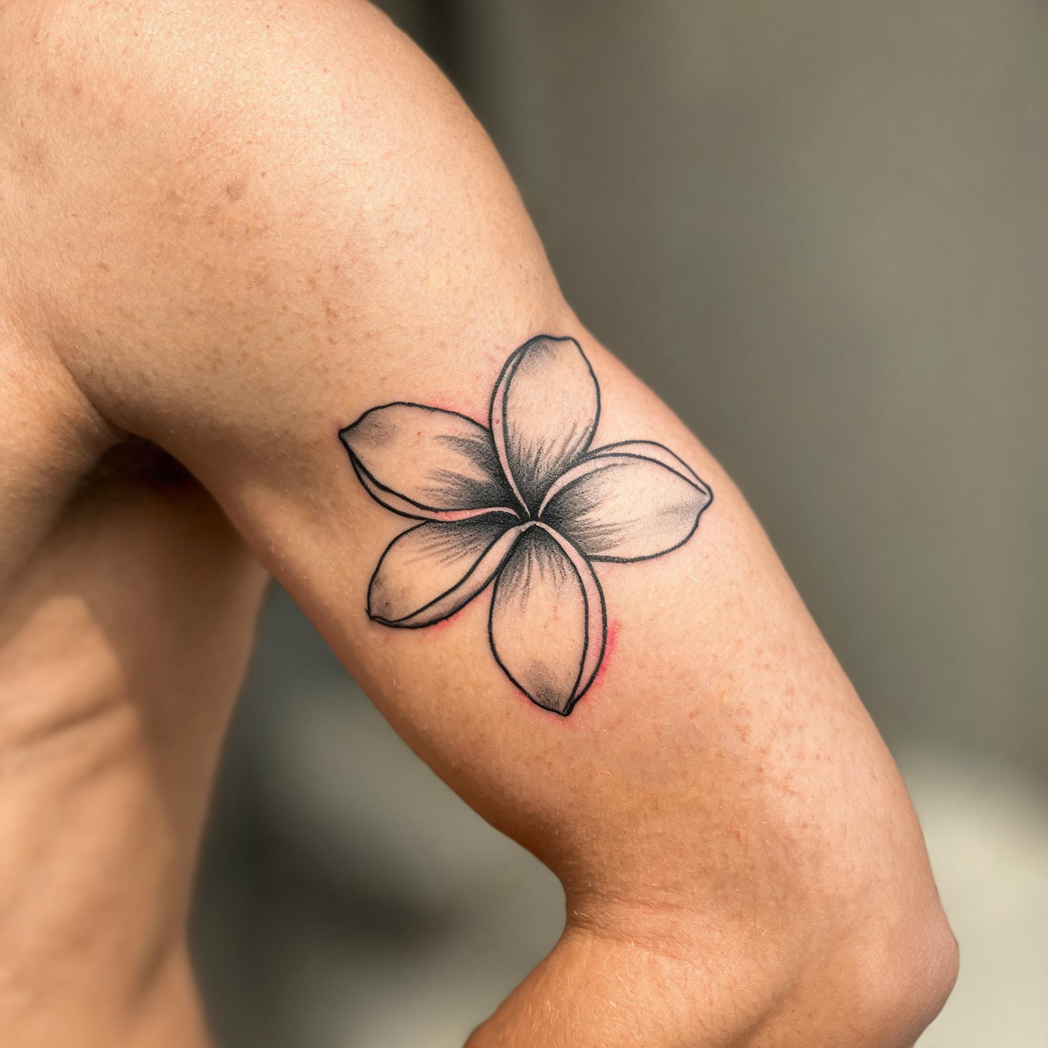A minimalist Hawaiian flower tattoo image on the outer upper arm, shown in black and grey ink against a clean neutral background.