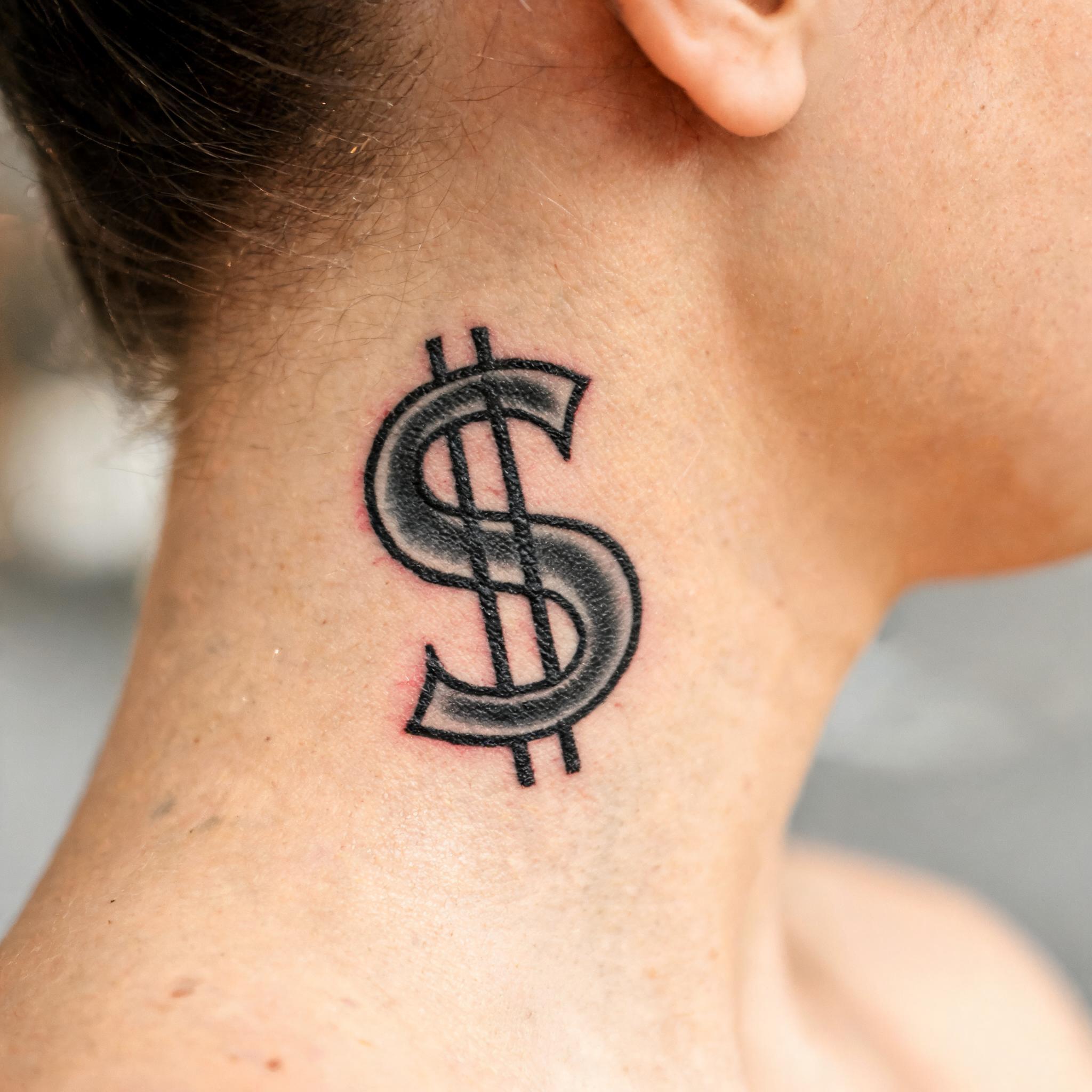 A neo-traditional dollar sign tattoo image on the nape of the neck, shown in black and grey on a white background.