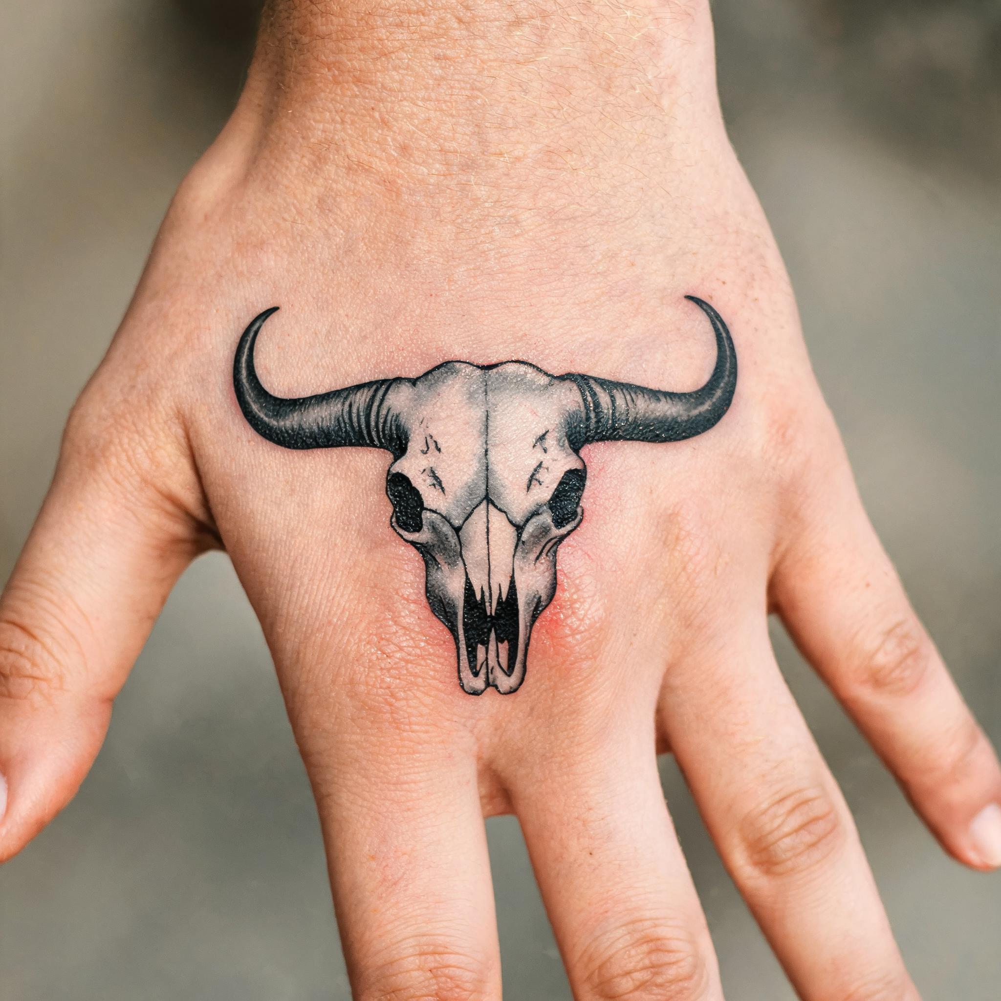 A blackwork bull skull tattoo image on the back of a hand, shown in high-contrast black and grey ink against a clean neutral background.