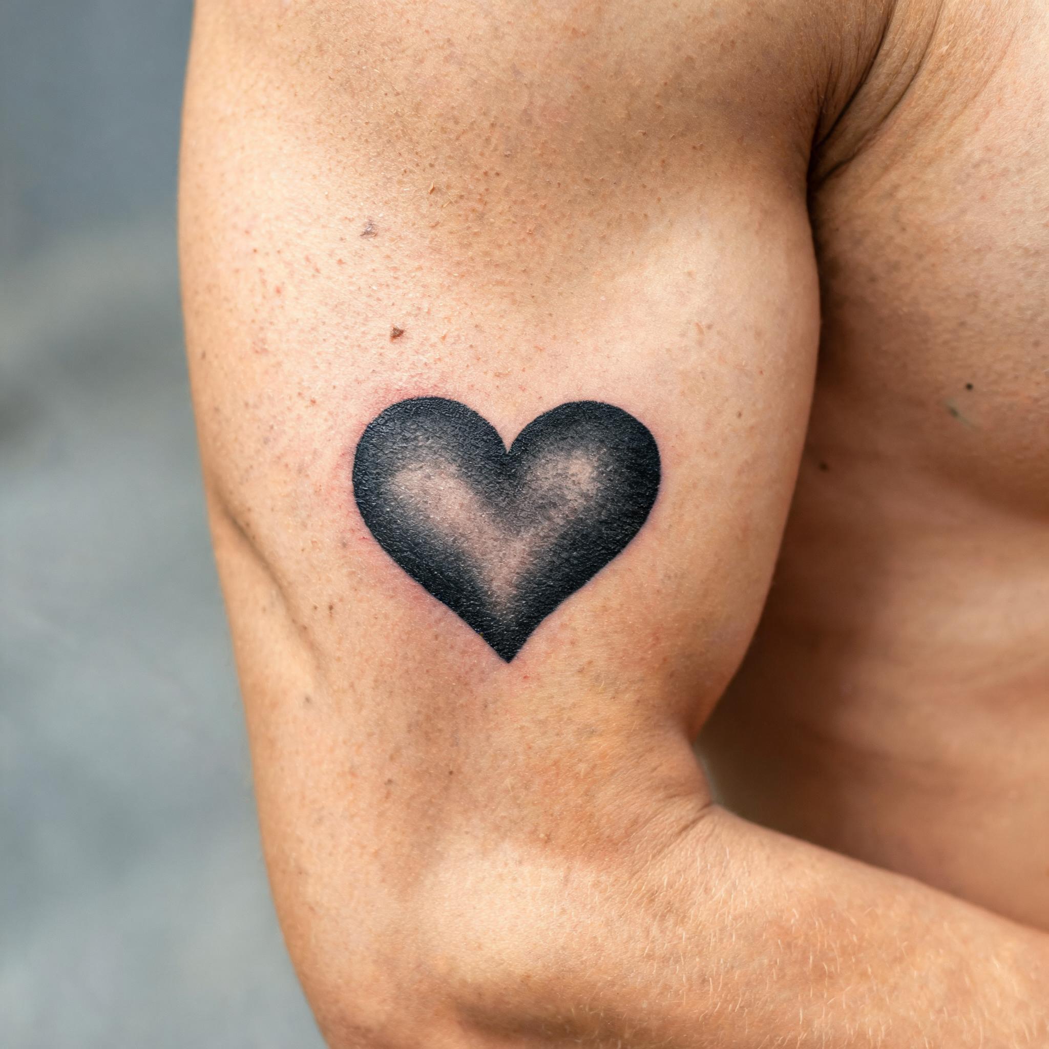 Blackwork heart tattoo images in solid black and greywash, shown on the outer upper arm against a clean neutral background.