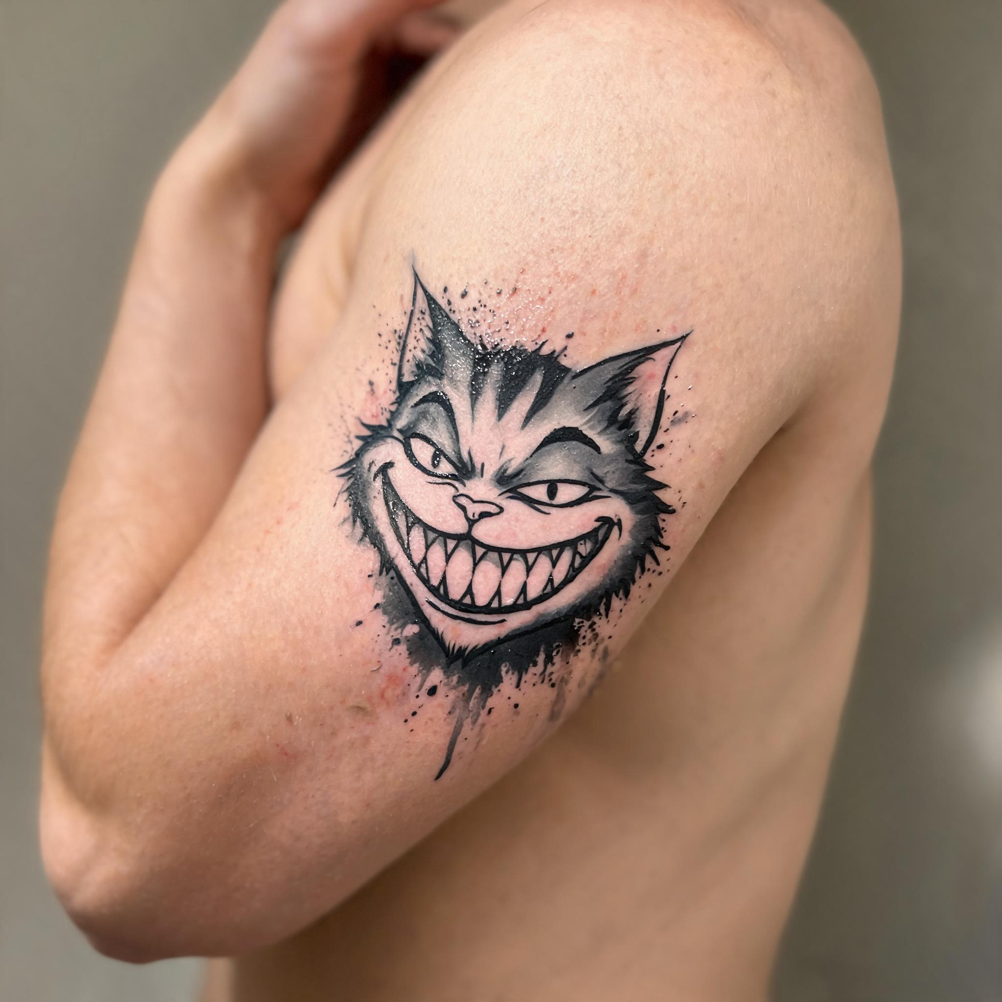 Black and grey trash polka Cheshire cat tattoo image on an inner bicep, shown on white background.