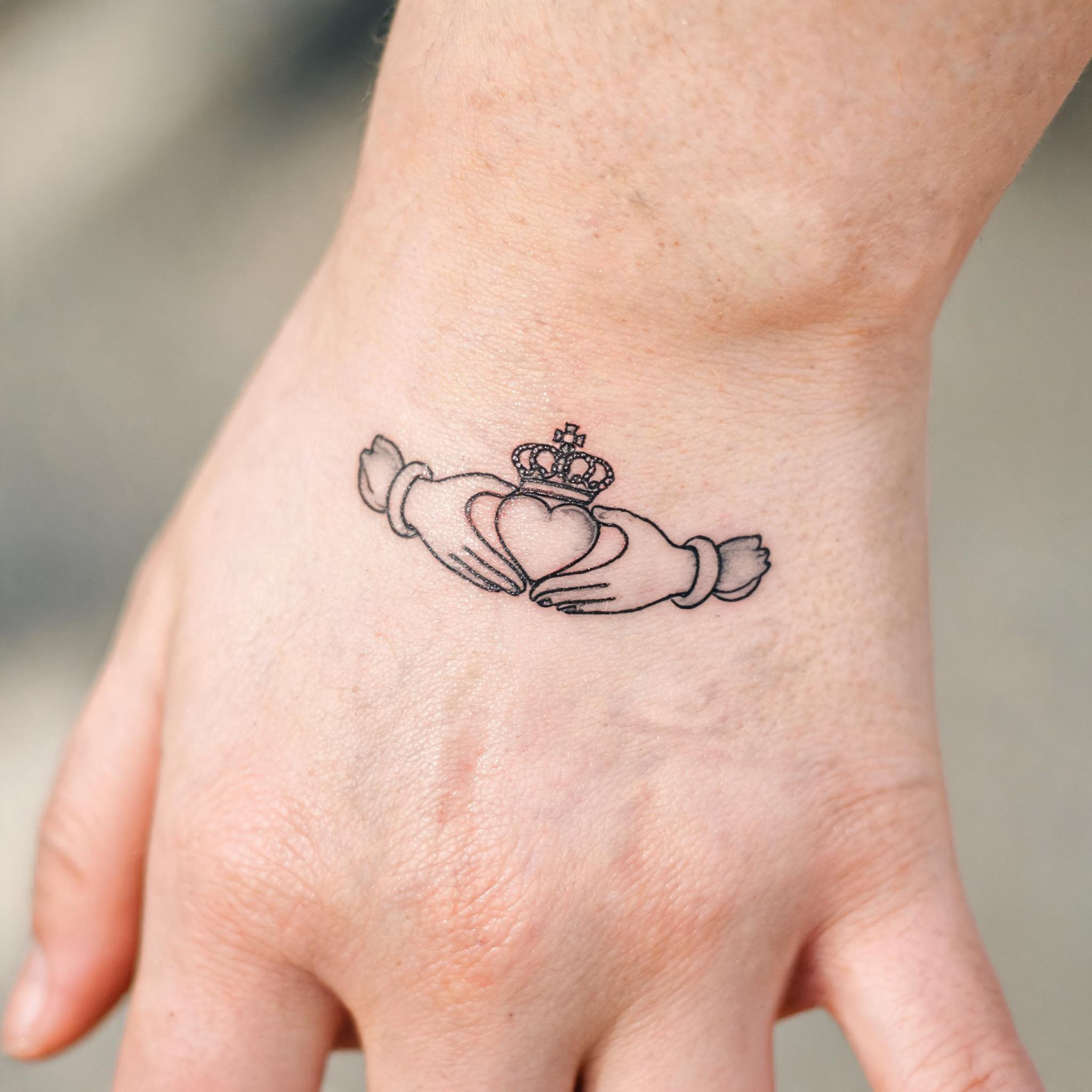 Fine line Claddagh tattoo on a wrist, black and grey with delicate single-needle lines on a clean neutral background, tattoo image.