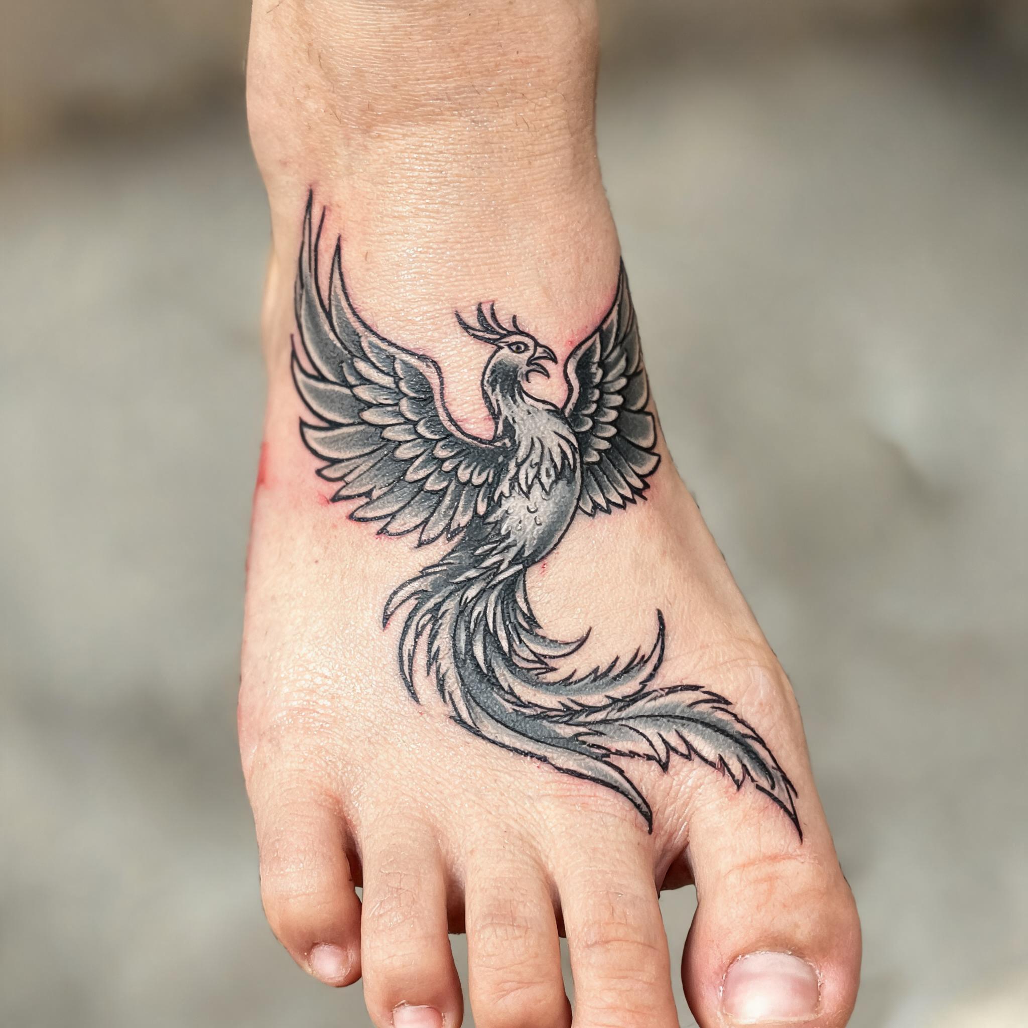 Black and grey American Traditional tattoo image of a phoenix bird on the top of a foot, shown on a clean neutral background.