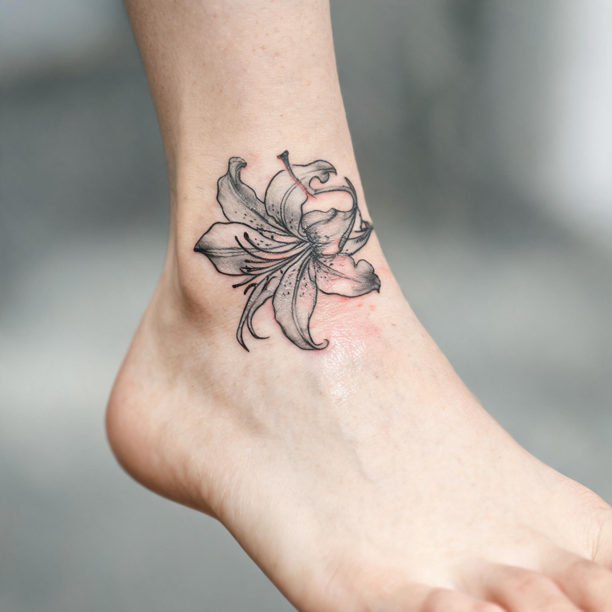 Minimalist black-and-grey tiger lily tattoo on an ankle, shown on skin against a clean neutral background as a tattoo image.