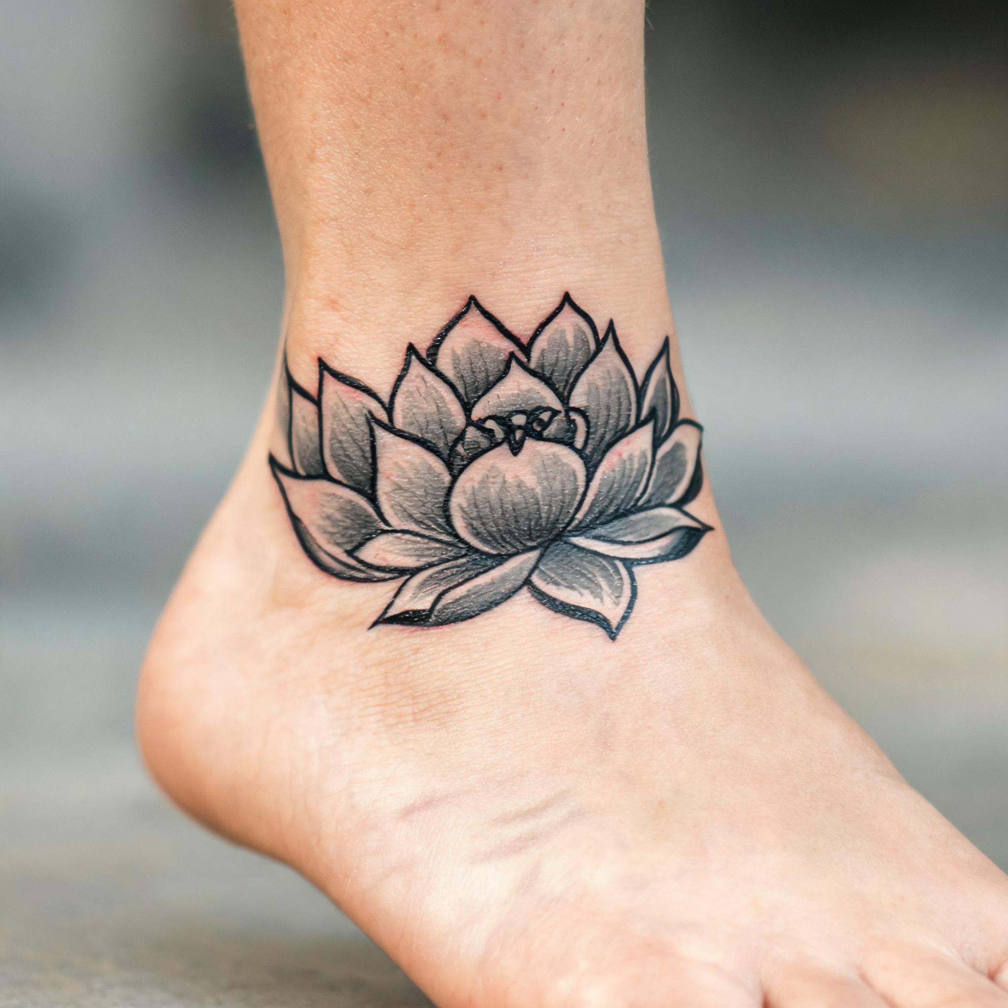 American traditional lotus tattoo images on an ankle, bold black outlines and greywash shading, shown on skin in a white-background tattoo photo.