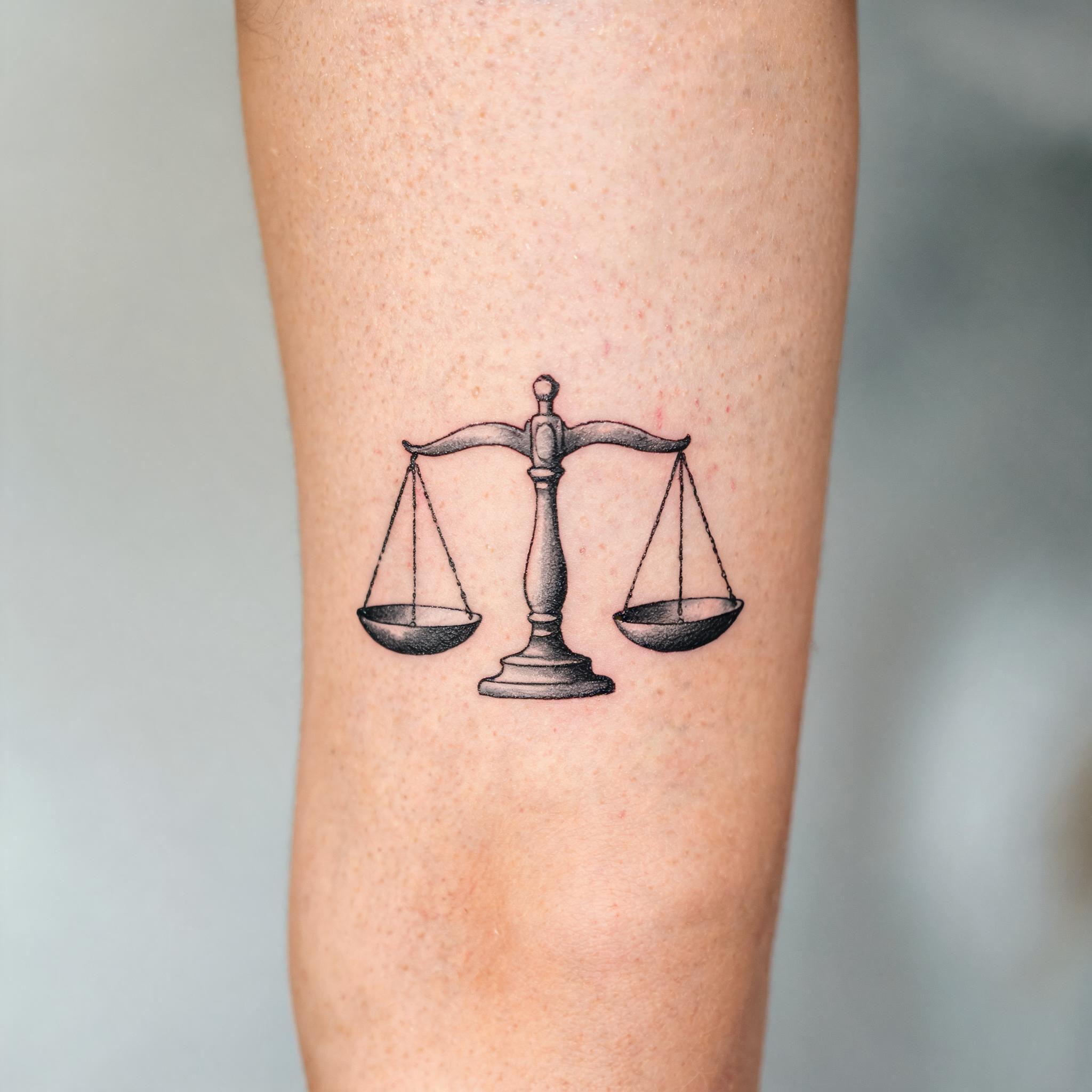 A black and grey Libra scale tattoo image on the front thigh, shown as realistic tattoo artwork on skin against a neutral background.