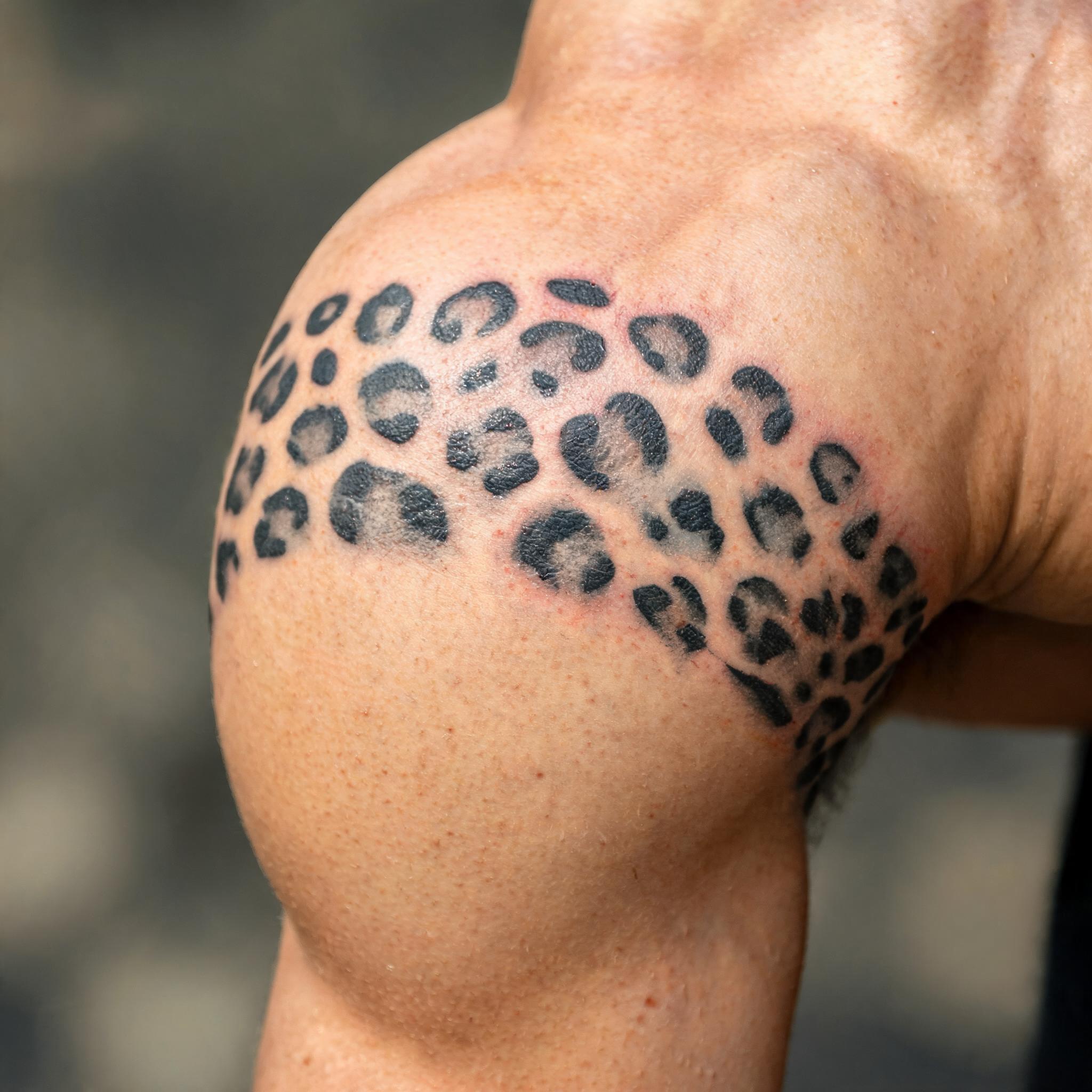 Minimalist black and grey leopard print tattoo on the outer upper arm, shown on a clean neutral background as a tattoo image.