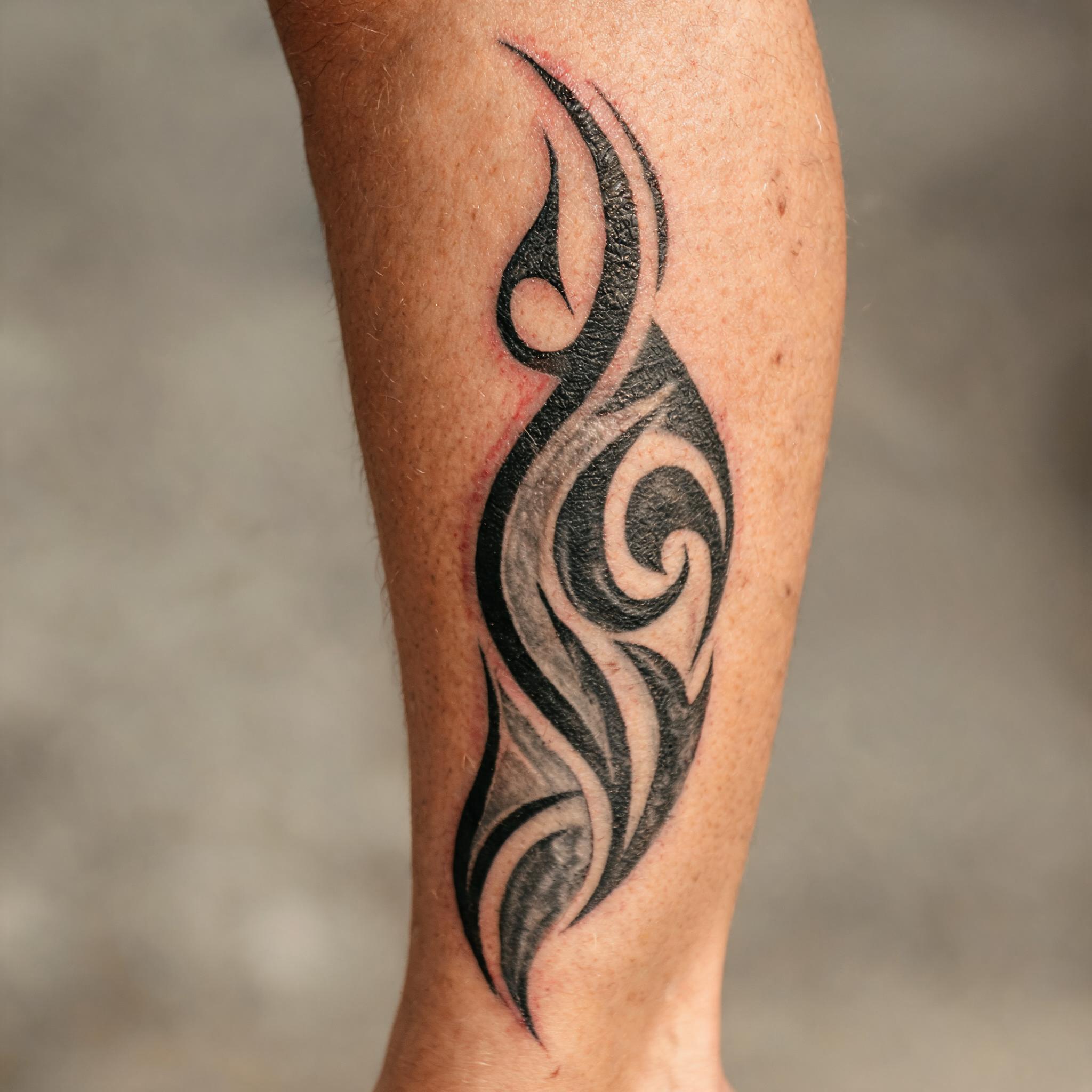 A black and grey tribal sleeve tattoo image running down the calf on a person's lower leg, shown on a clean neutral background.