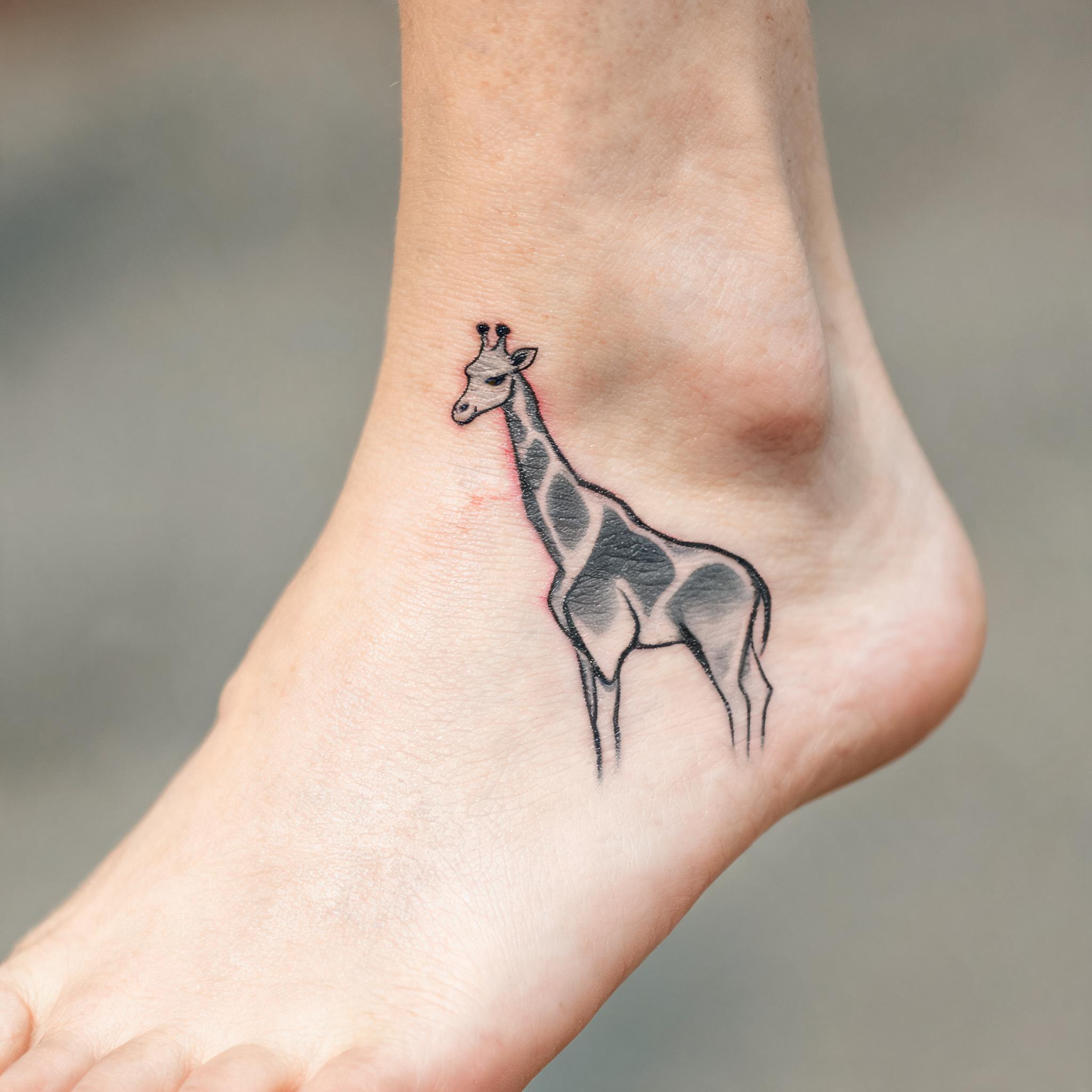 A minimalist giraffe tattoo image design inked on an ankle, shown in black and grey linework on a plain background as a tattoo image.
