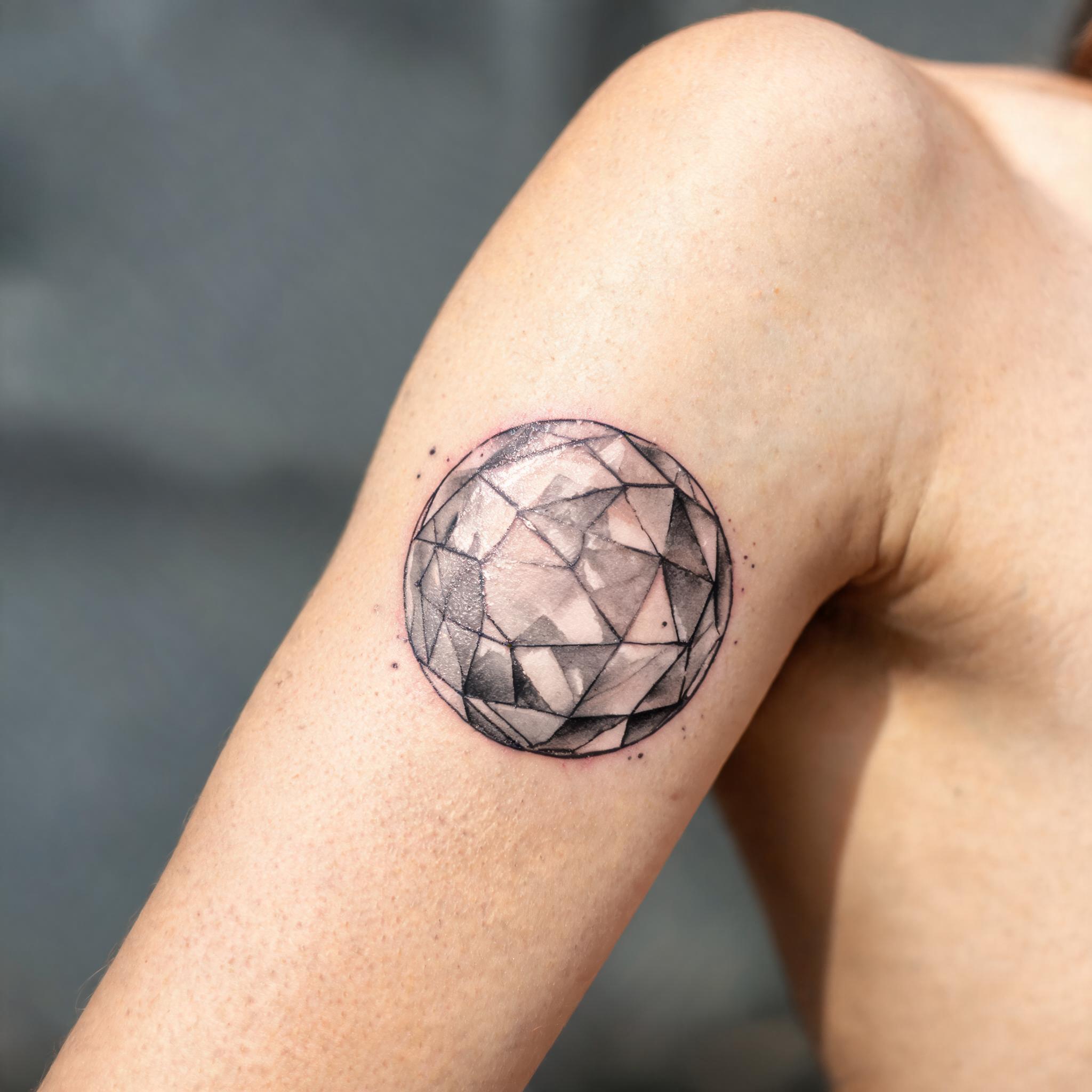 A black-and-grey watercolor tattoo image of a crystal ball design on the inner bicep, shown on skin against a plain neutral background.