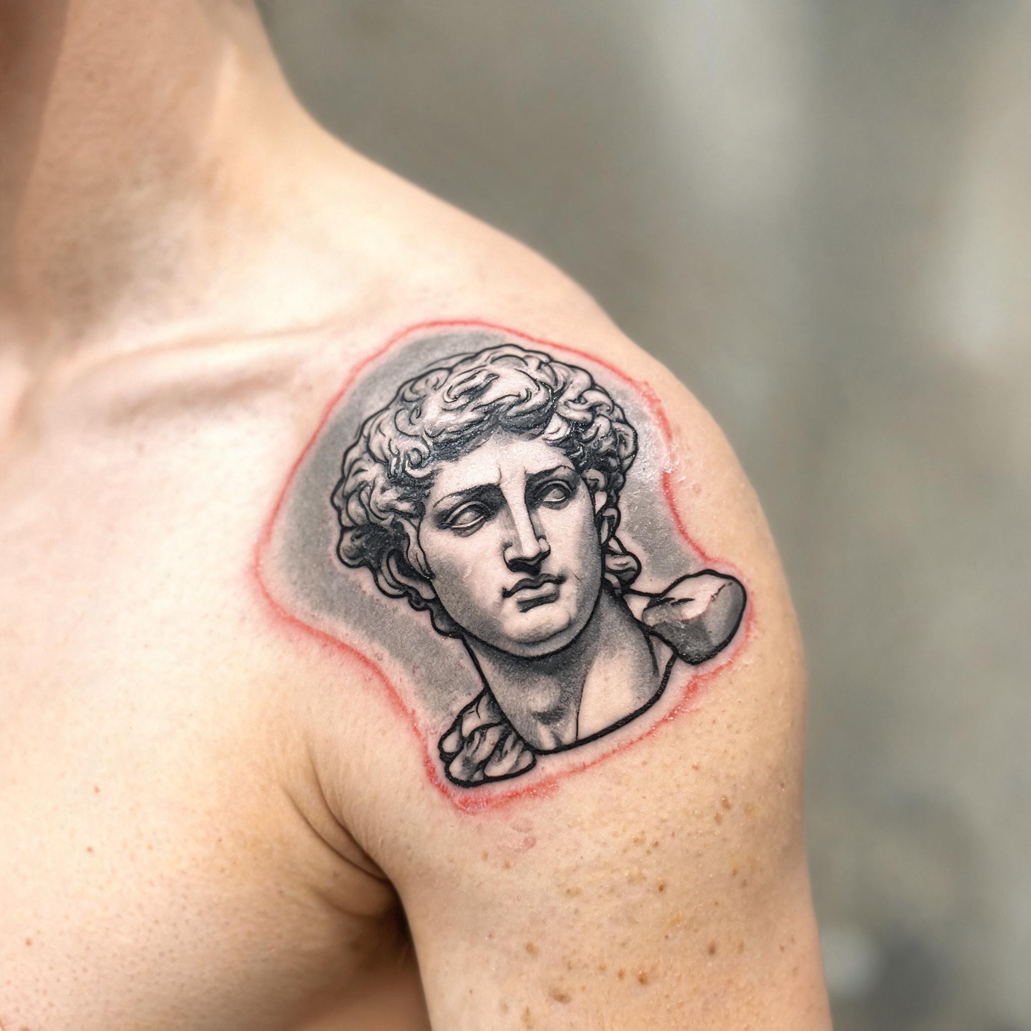 Black and grey American Traditional Greek statue tattoo image design on a shoulder cap, shown on skin against a neutral background.