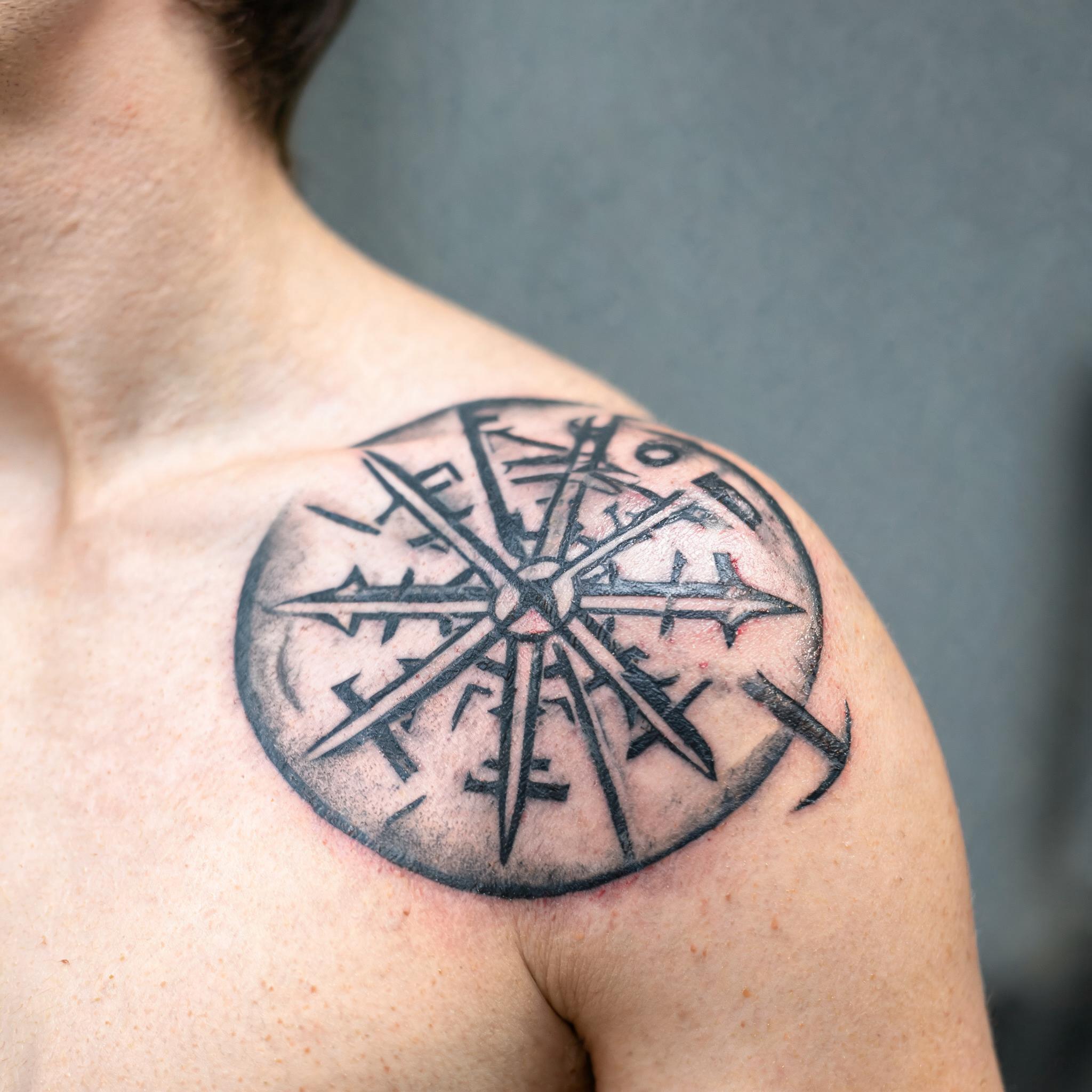 A viking compass tattoo design in black and grey realism, photographed on a shoulder cap against a clean neutral background as a tattoo image.