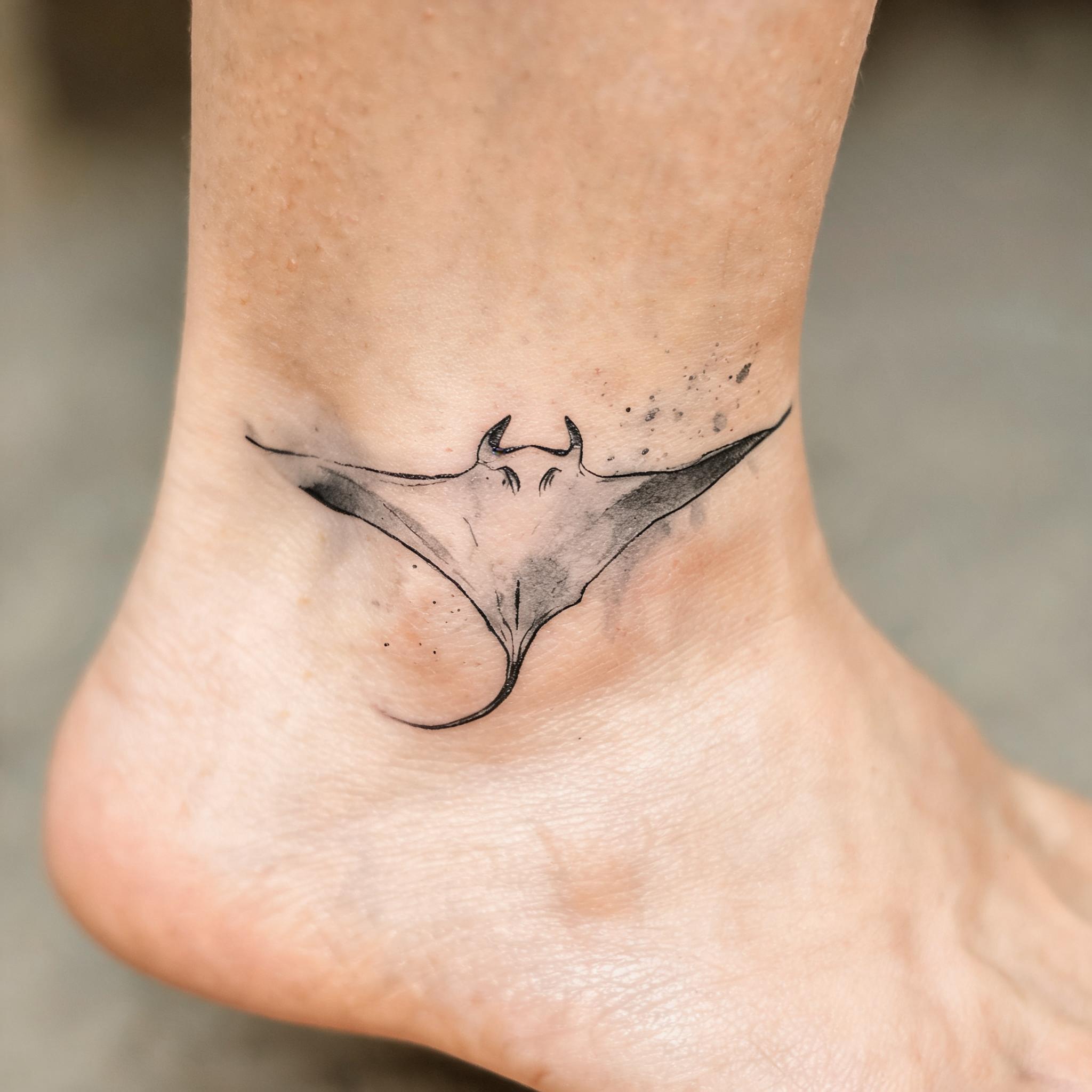 A black and grey watercolor manta ray ocean tattoo design on an ankle, shown as a tattoo image on clean white background.