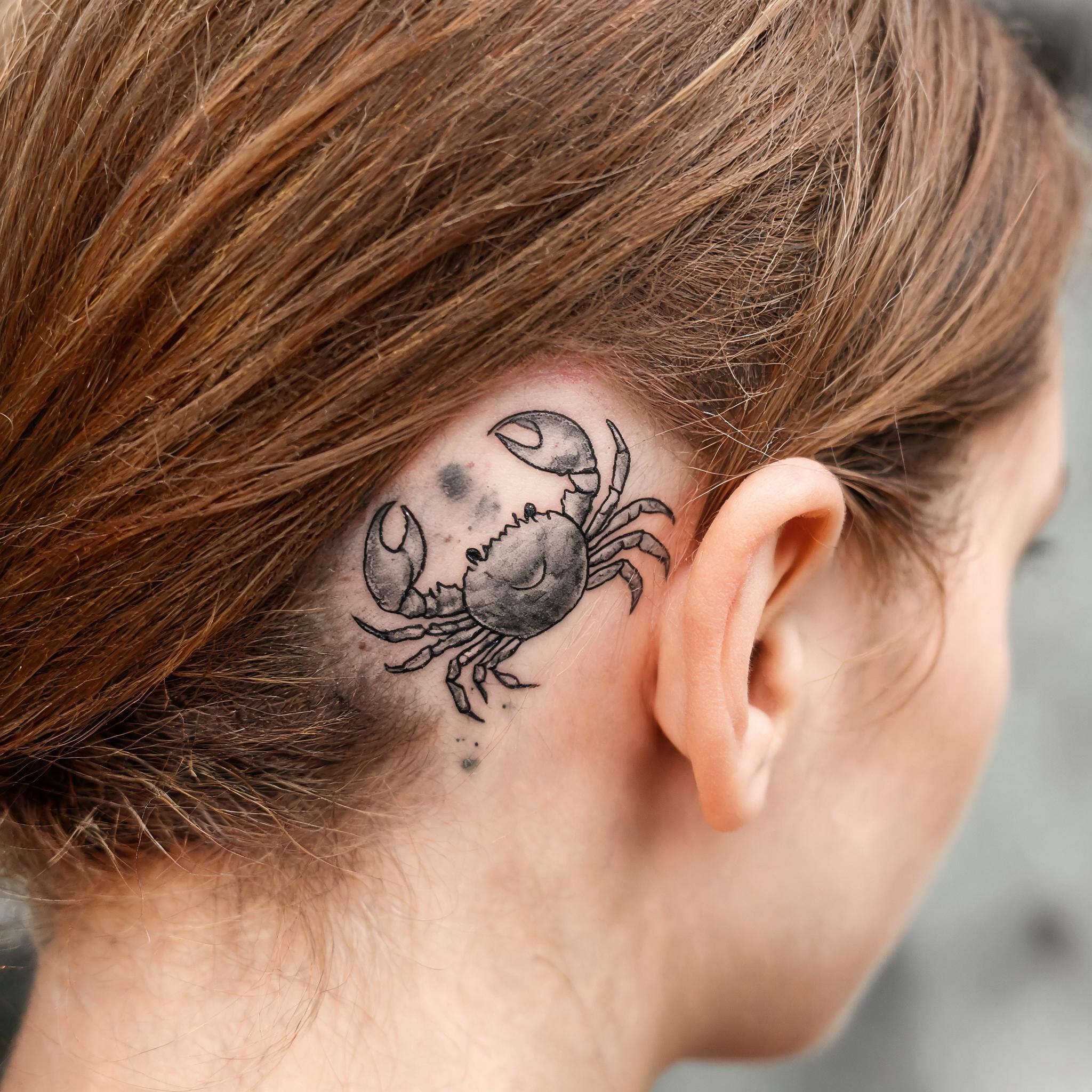 Watercolor cancer crab zodiac tattoo design in black and grey ink behind the ear, shown on skin against a clean neutral background.