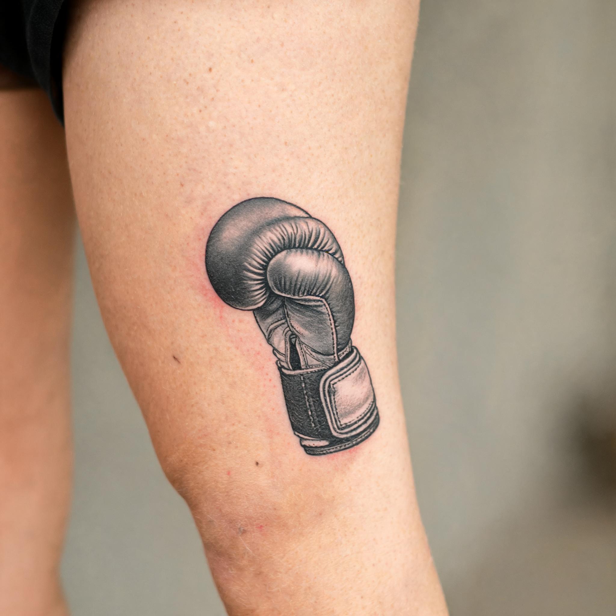 Black and grey realistic boxing gloves tattoo design on an outer thigh, shown on skin in a tattoo photo.