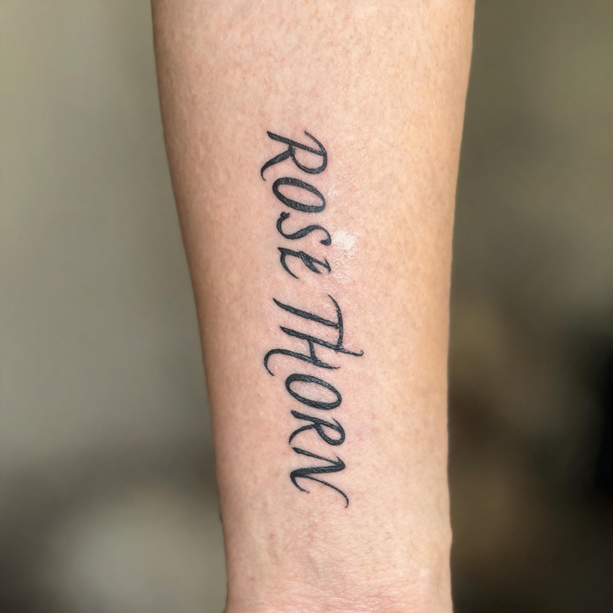 A black and grey single rose thorn tattoo design in elegant calligraphy script on an outer forearm, shown on a white background as a tattoo image.