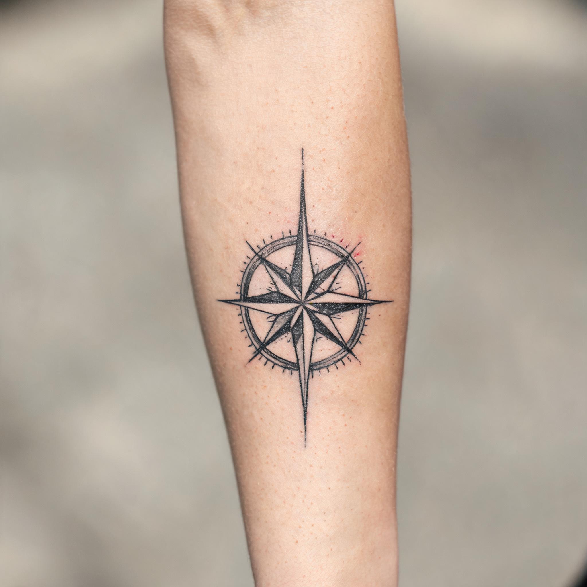 Black and grey illustrative compass rose tattoo on a person's outer forearm, shown on real skin against a neutral background.