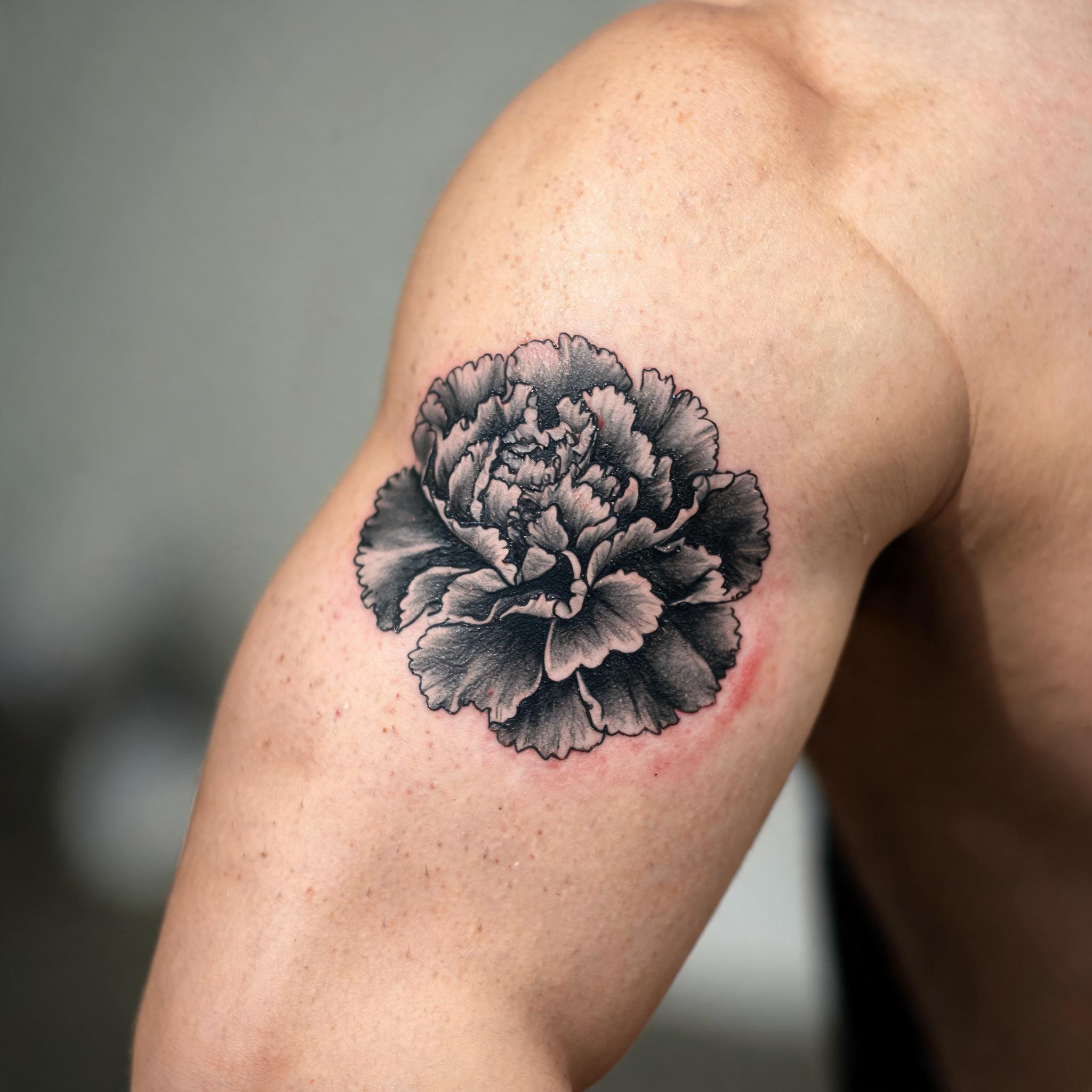 Blackwork carnation flower tattoo image design on the outer upper arm, shown in a close-up photo on a clean neutral background.