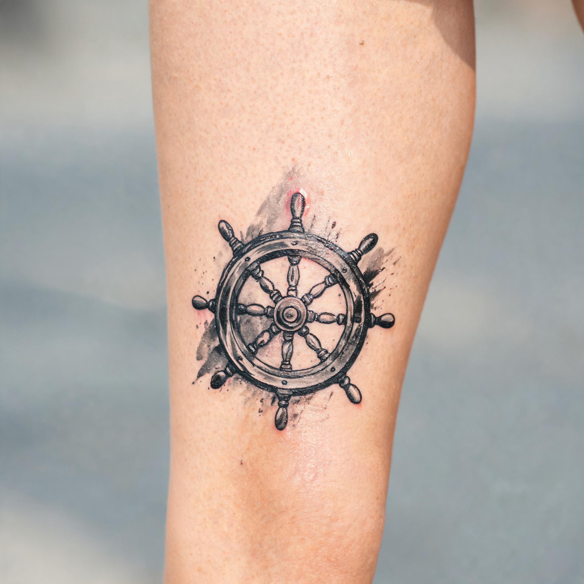 A black and grey trash polka tattoo image of a ship wheel on the outer thigh, shown on skin against a neutral background.
