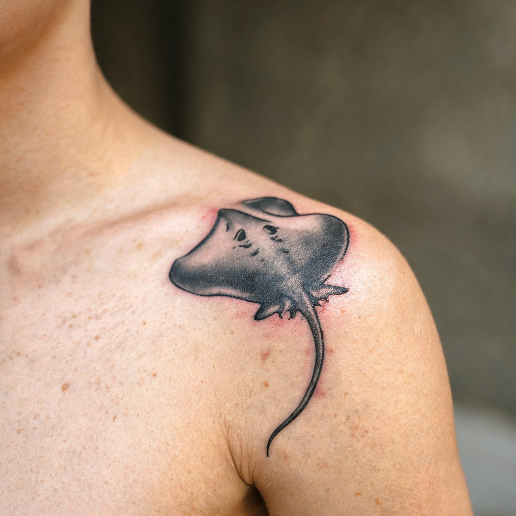 Black and grey stingray tattoo image on a shoulder cap, photorealistic close-up on white background showing shaded detail.