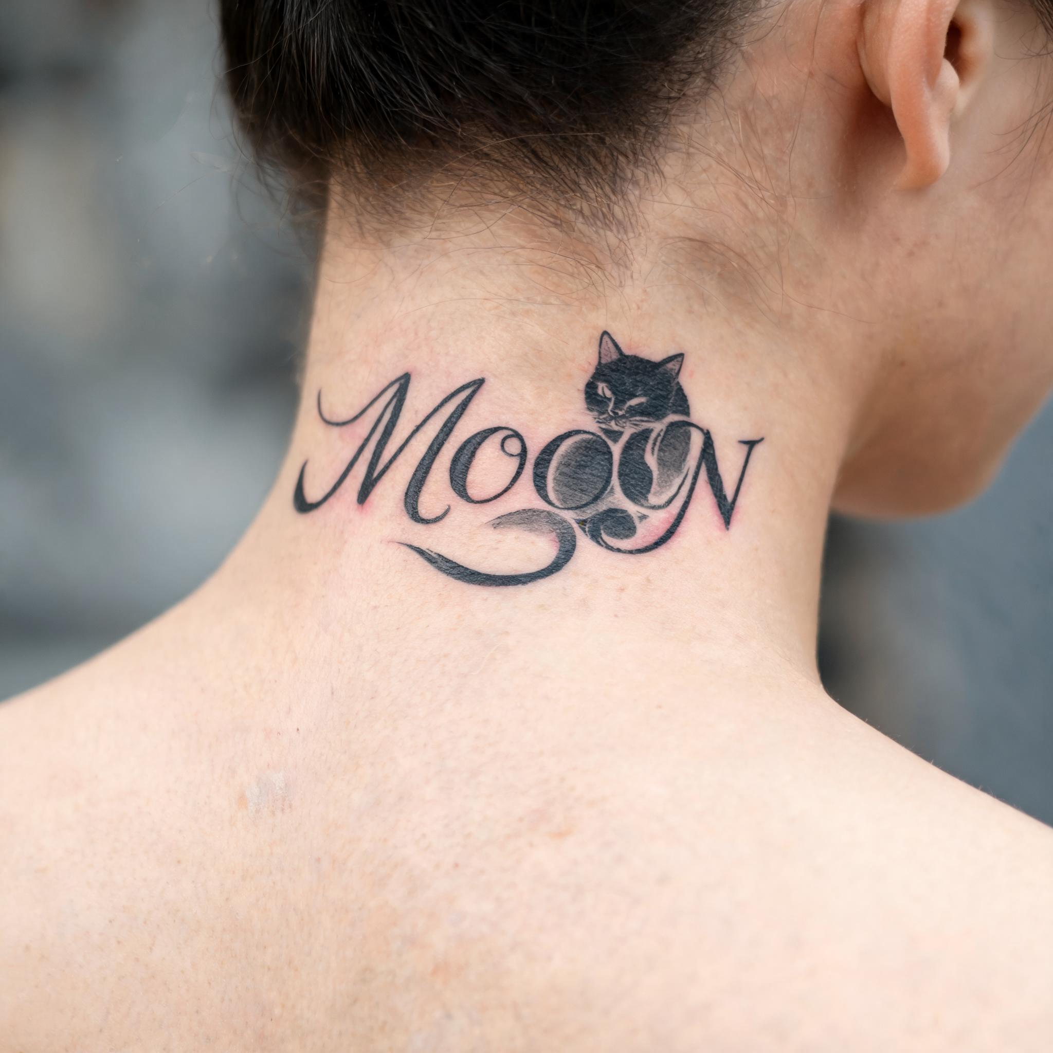 Black cat moon tattoo design on the nape of the neck, shown in black and grey script lettering on a person's skin.