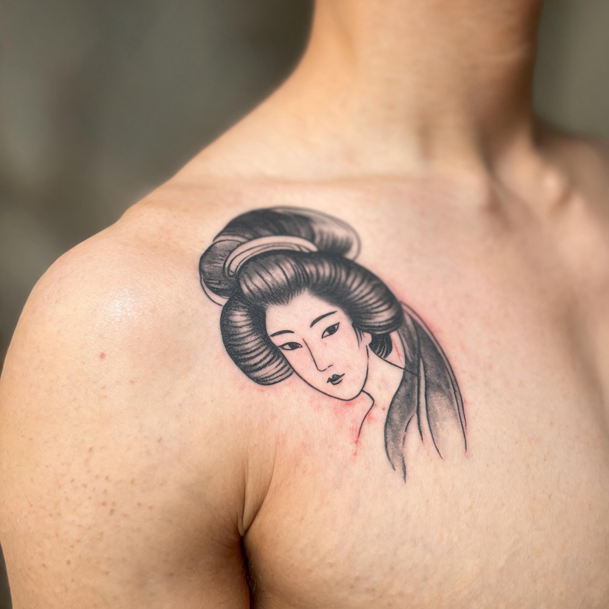 Minimalist black and grey geisha tattoo image on a shoulder cap, shown in a close-up photo against a plain background.