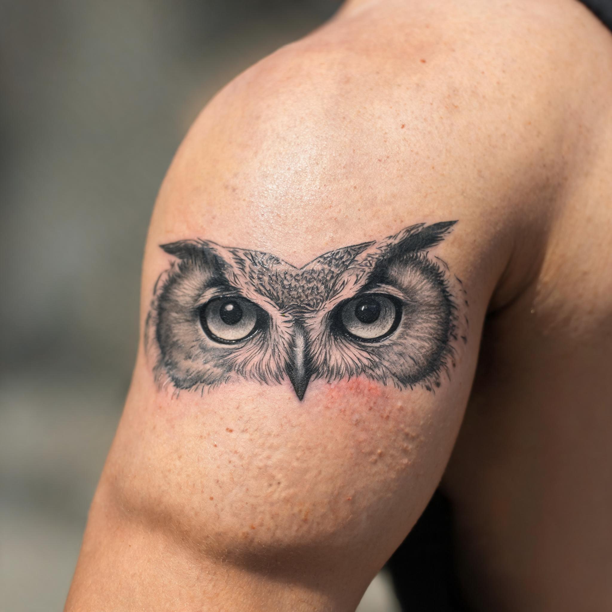 Illustrative owl eyes tattoo image design in black and grey ink on the outer upper arm, shown on skin against a clean neutral background.