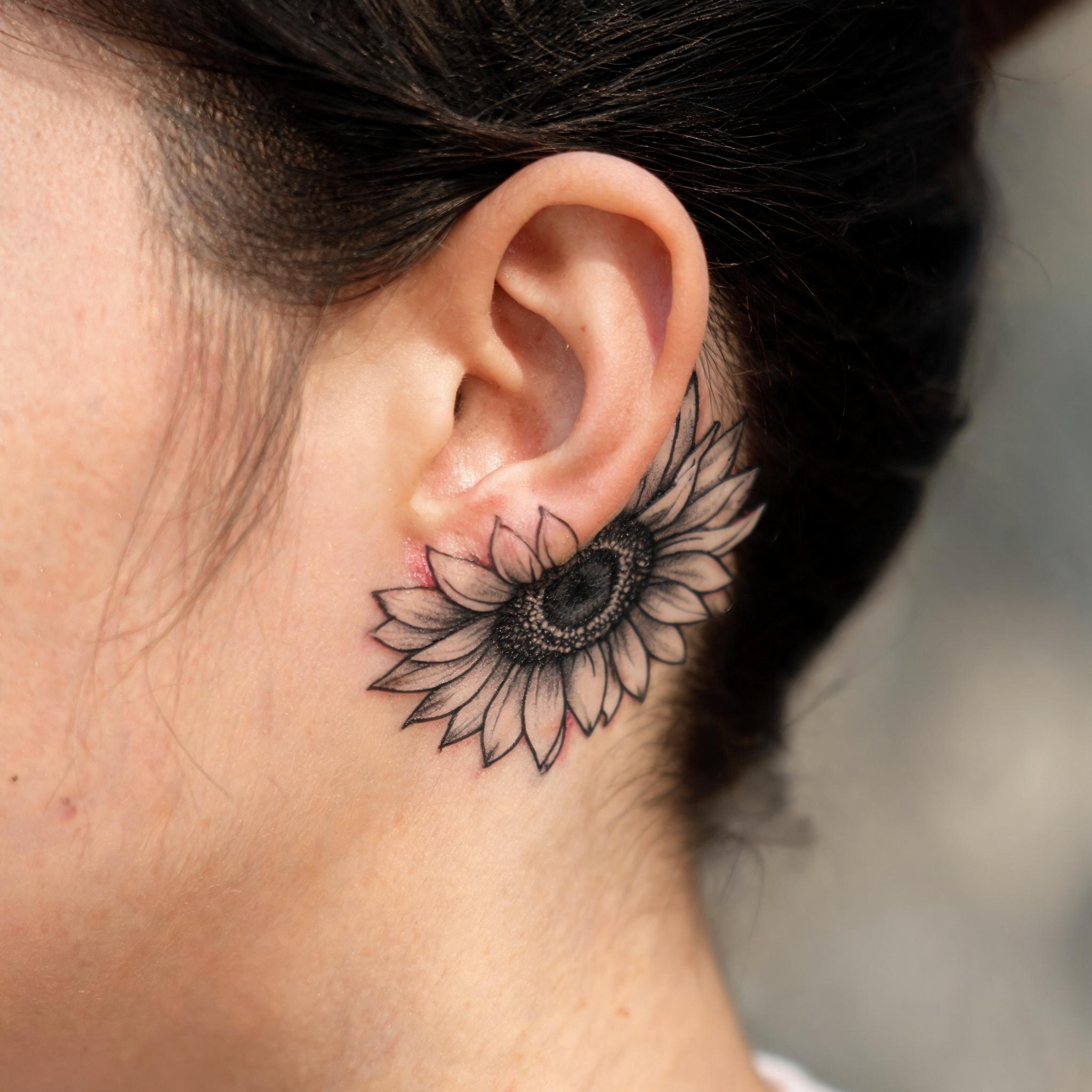 Illustrative black-and-grey sunflower tattoo design shown behind the ear, photographed on skin against a clean neutral background.