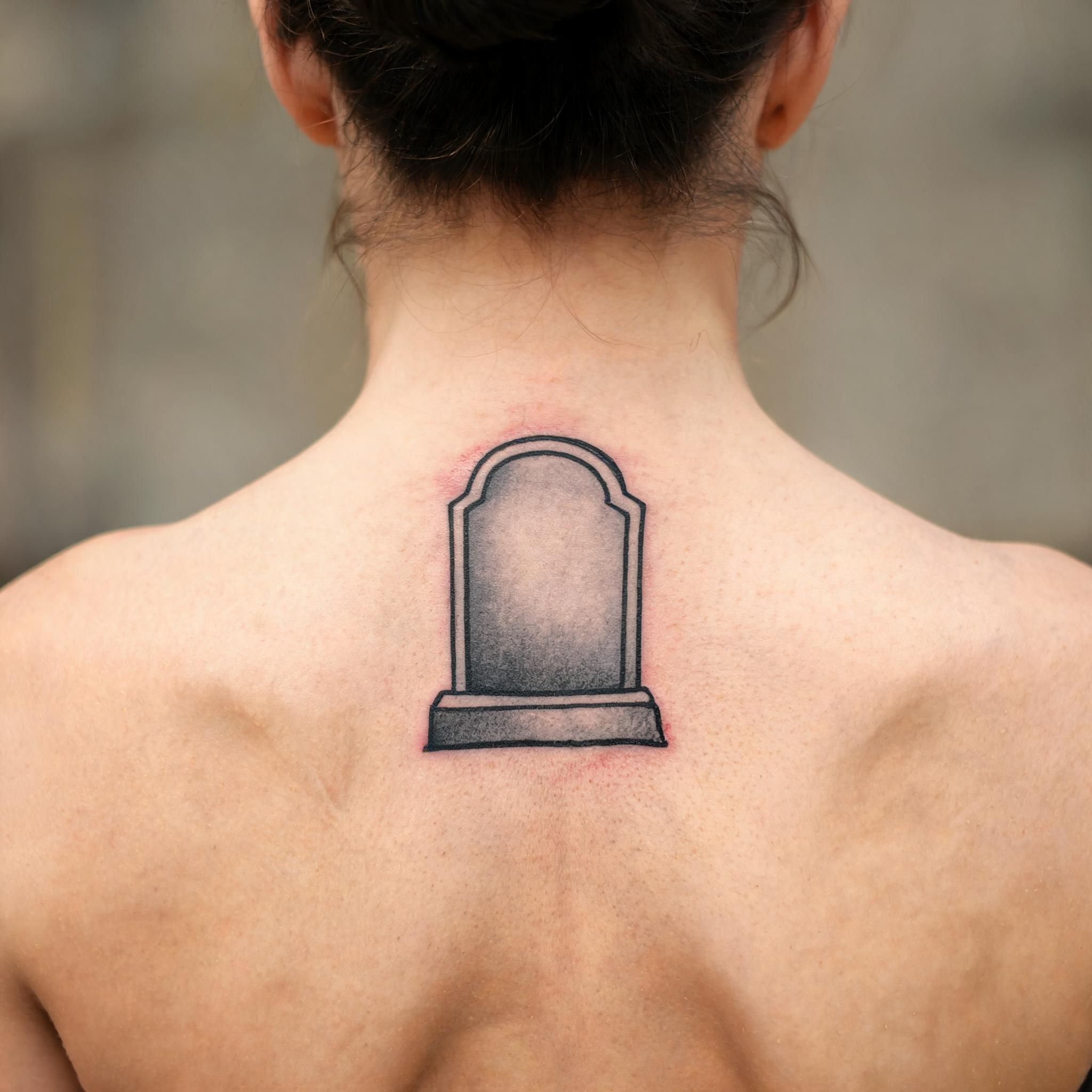 Black and grey American Traditional tombstone tattoo on an upper back, shown on skin against a neutral studio background as a tattoo image.