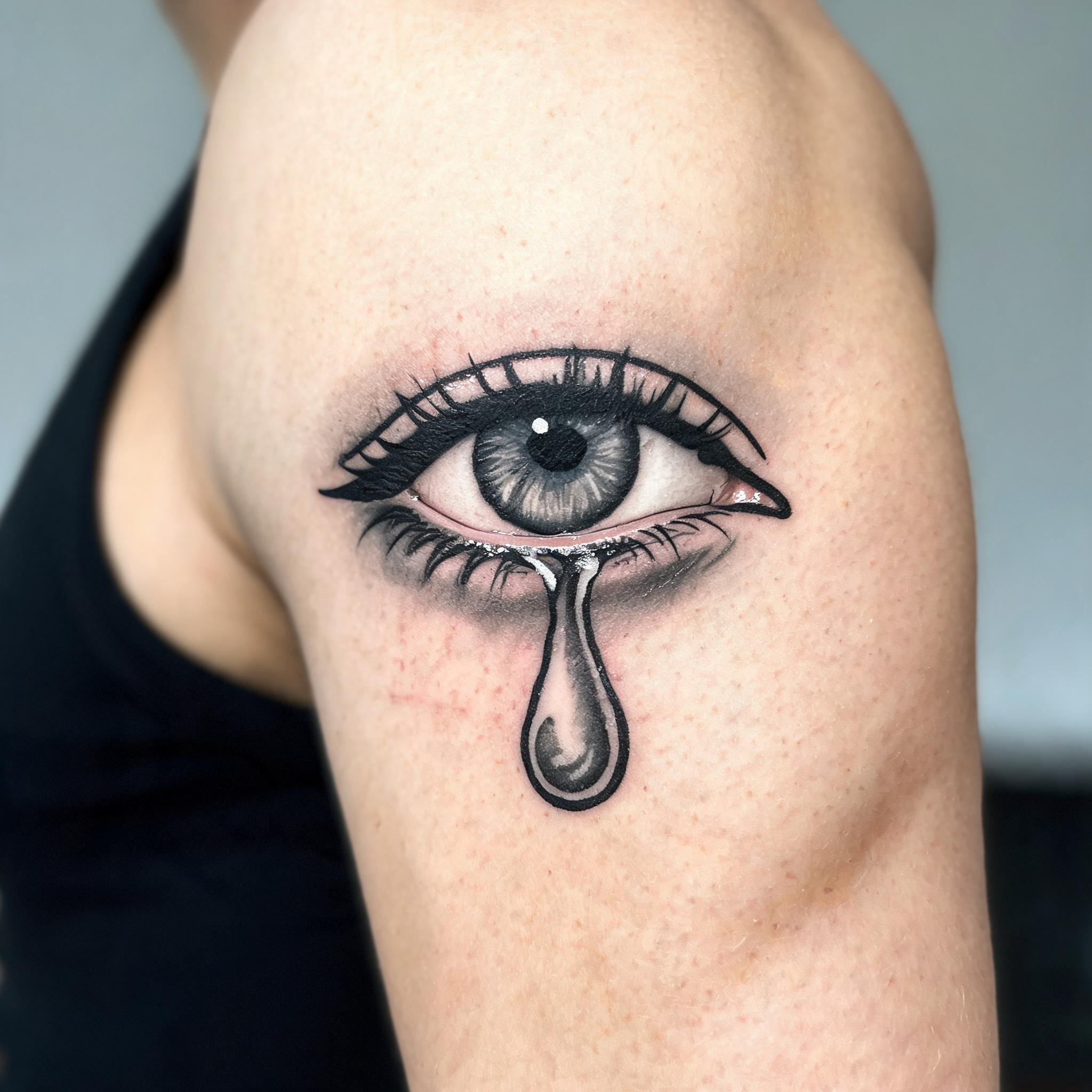 A crying eye tattoo design in American Traditional style on the outer upper arm, shown in black and grey ink on skin.
