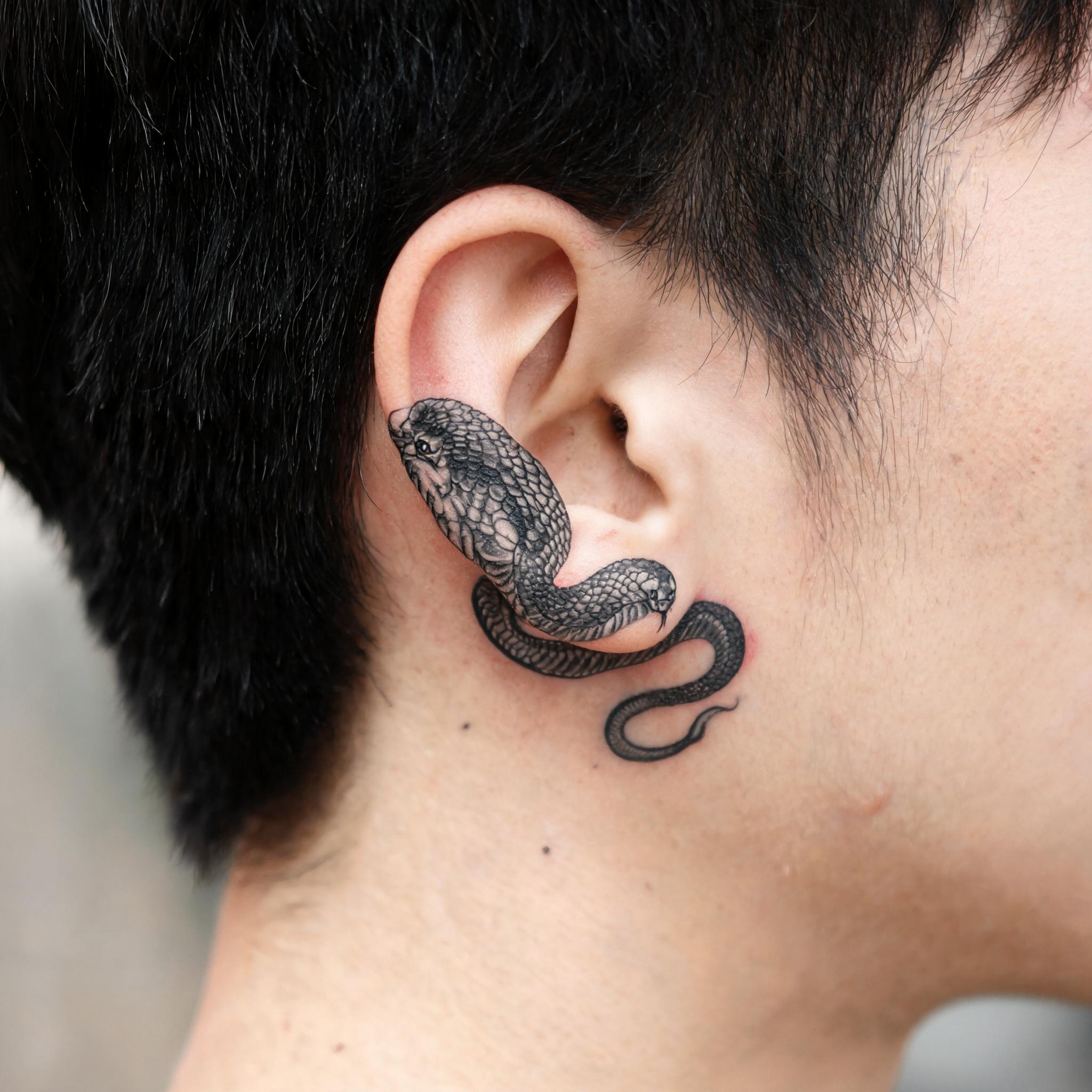 Black and grey king cobra tattoo image shown behind the ear, photorealistic close-up on skin with a neutral background.
