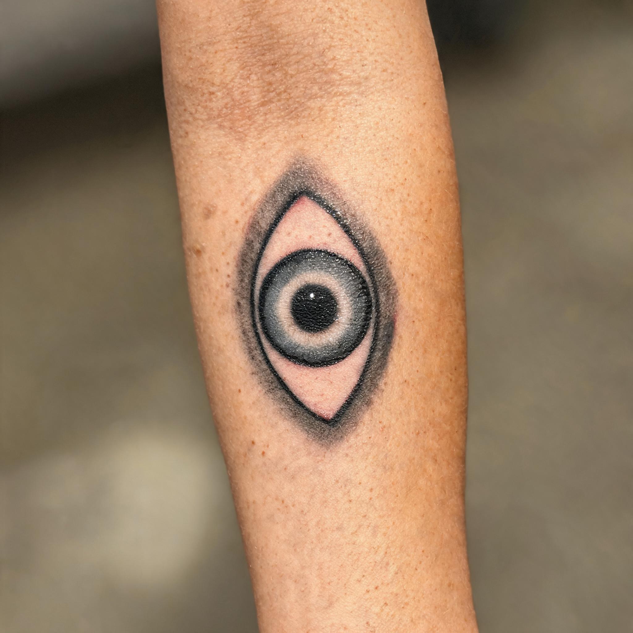 Black and grey evil eye protection tattoo on a person's inner forearm, shown in a realistic close-up tattoo photo against a neutral background.