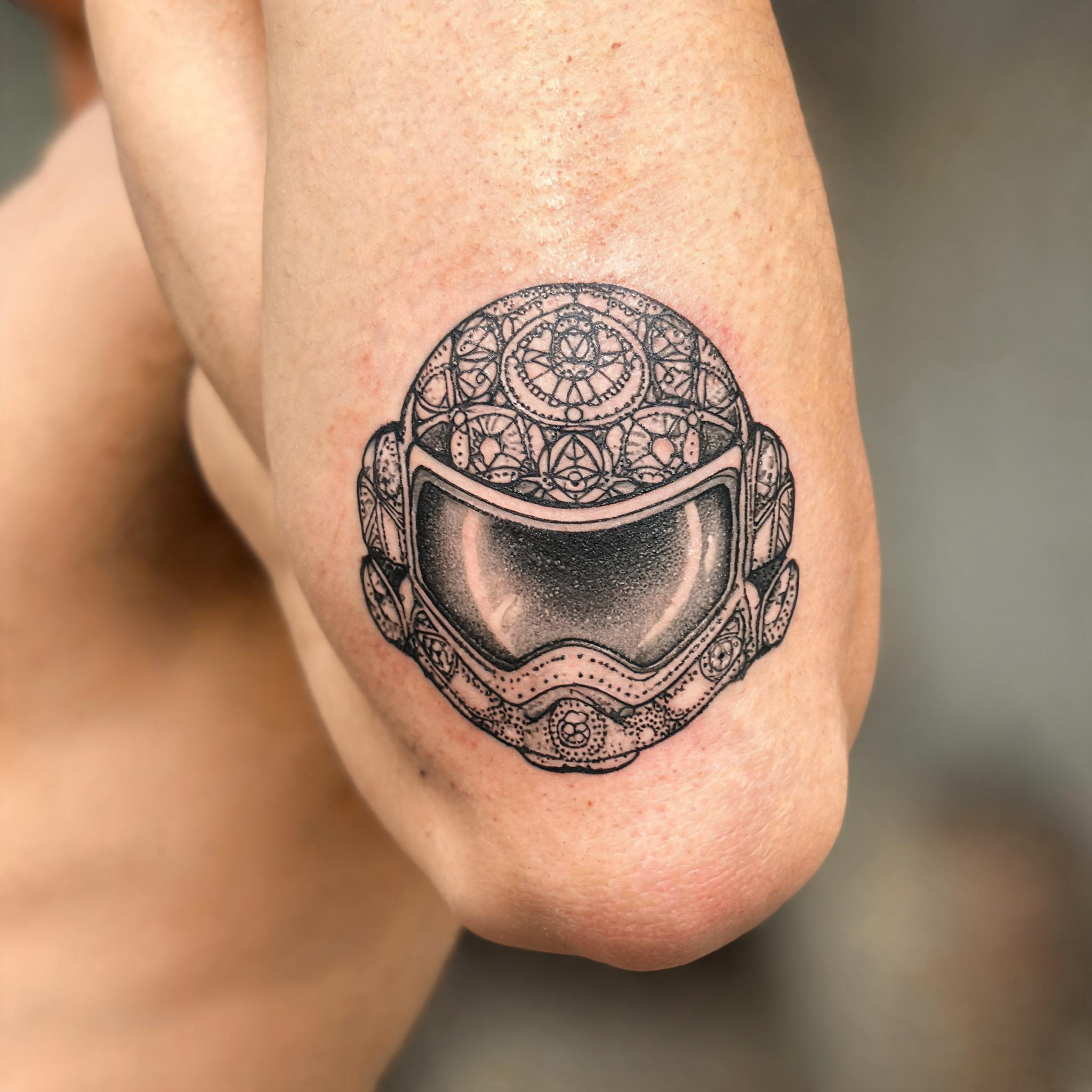 A black and grey astronaut helmet tattoo image design in geometric dotwork, shown on an inner bicep against a plain background.