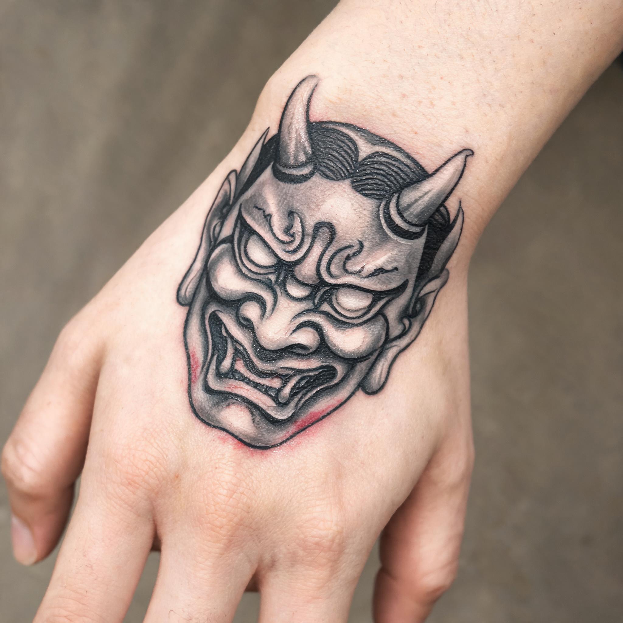 Hannya mask tattoo design in illustrative black and grey ink on a wrist, photographed on white background as a tattoo image.