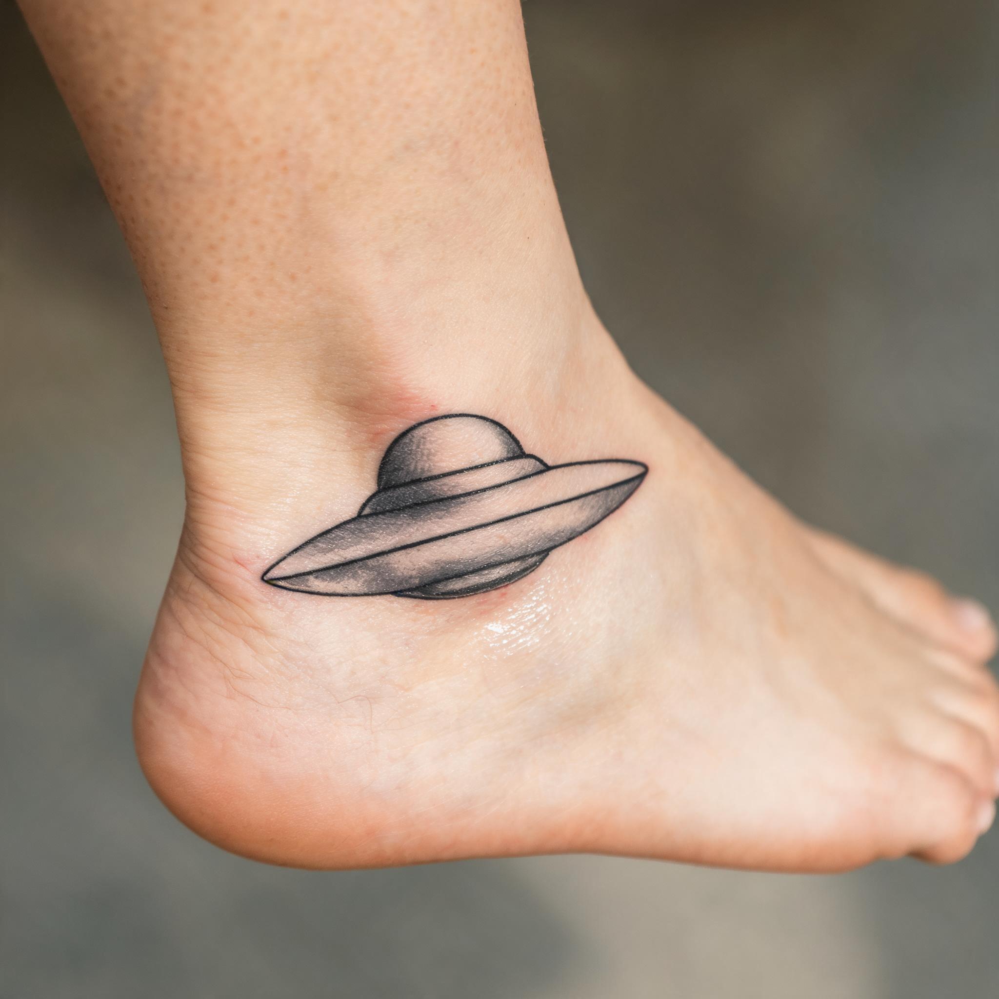 Minimalist alien UFO tattoo image design in black and grey ink on an ankle, photographed on skin against a neutral background.