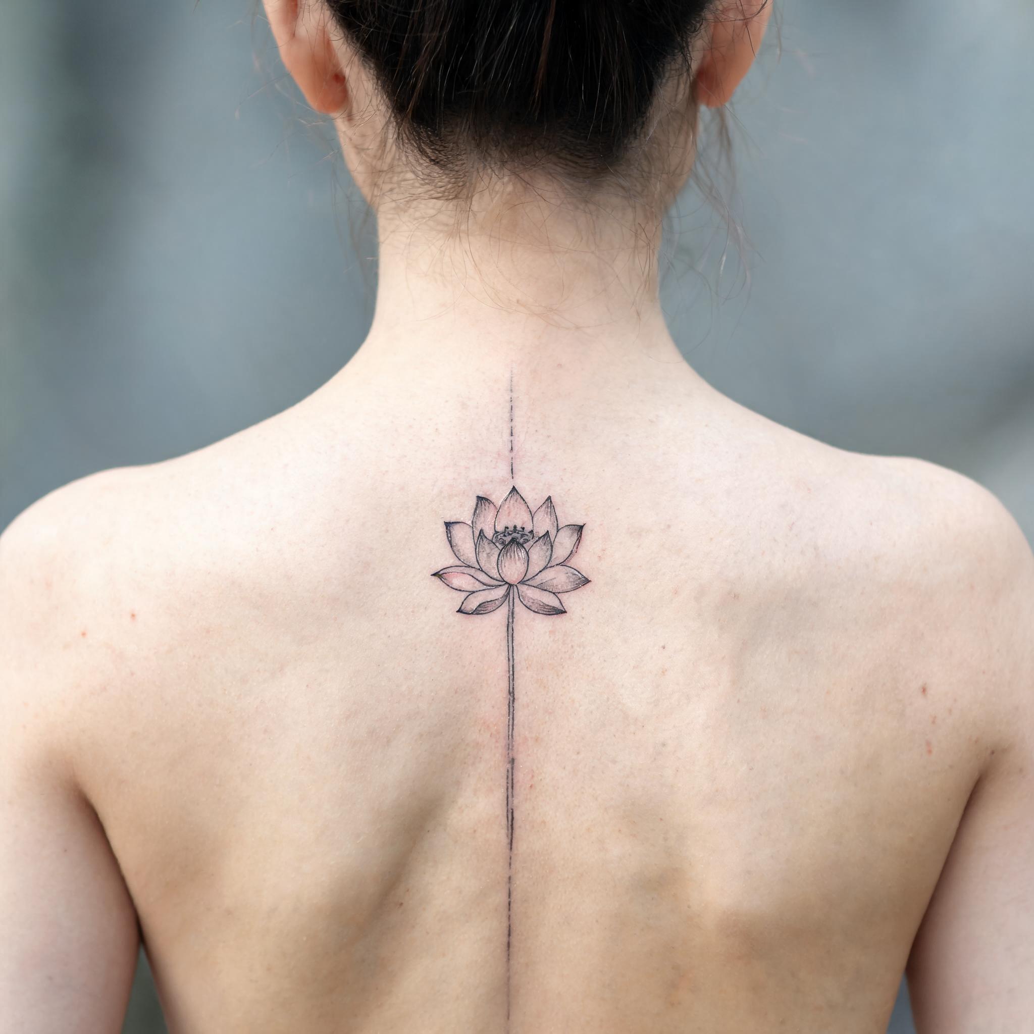 A minimal lotus flower tattoo design in fine line black and grey ink, shown on a person's spine against a plain background.