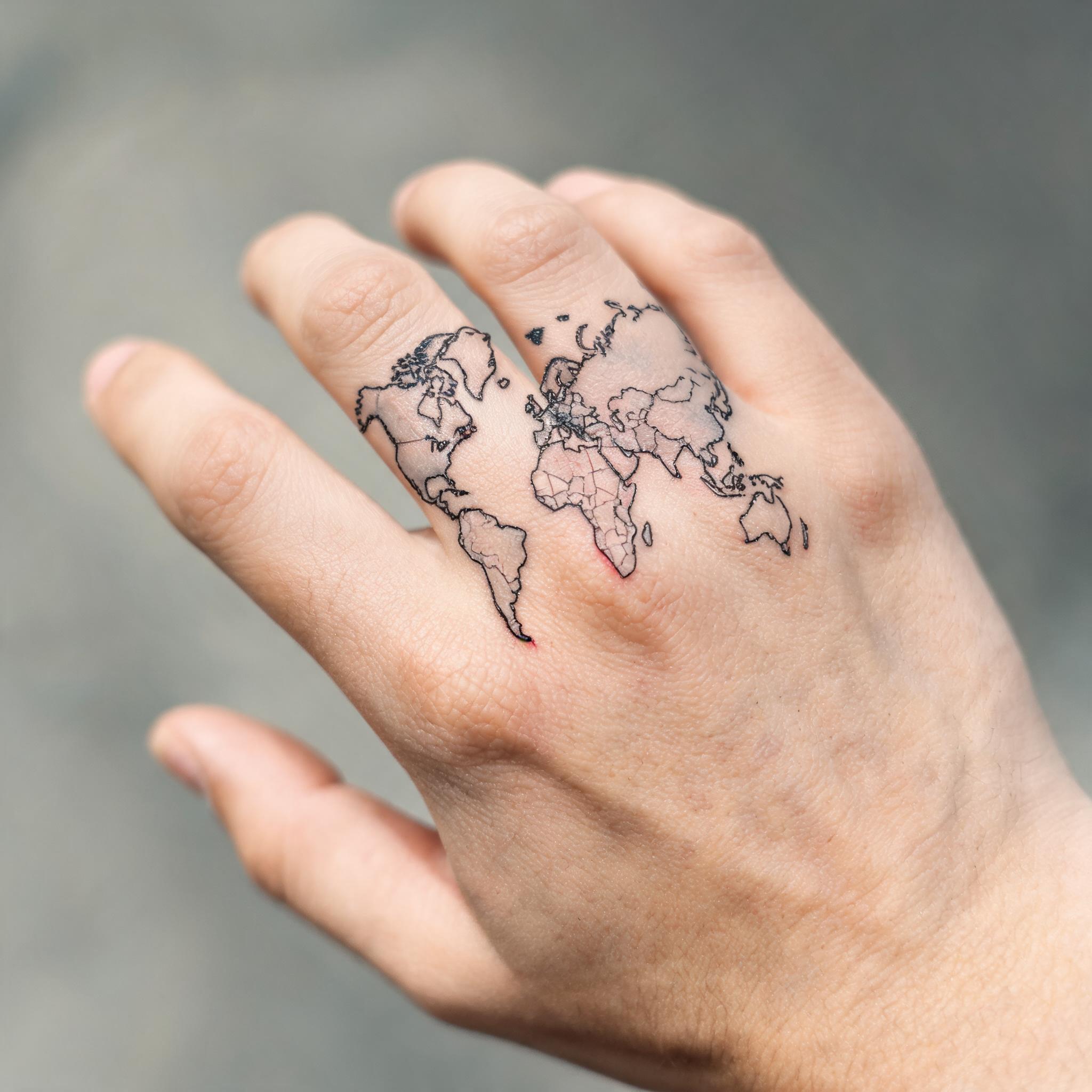 Fine line tattoo of Atlas holding the world, shown on a hand with fingers extended against a neutral background.