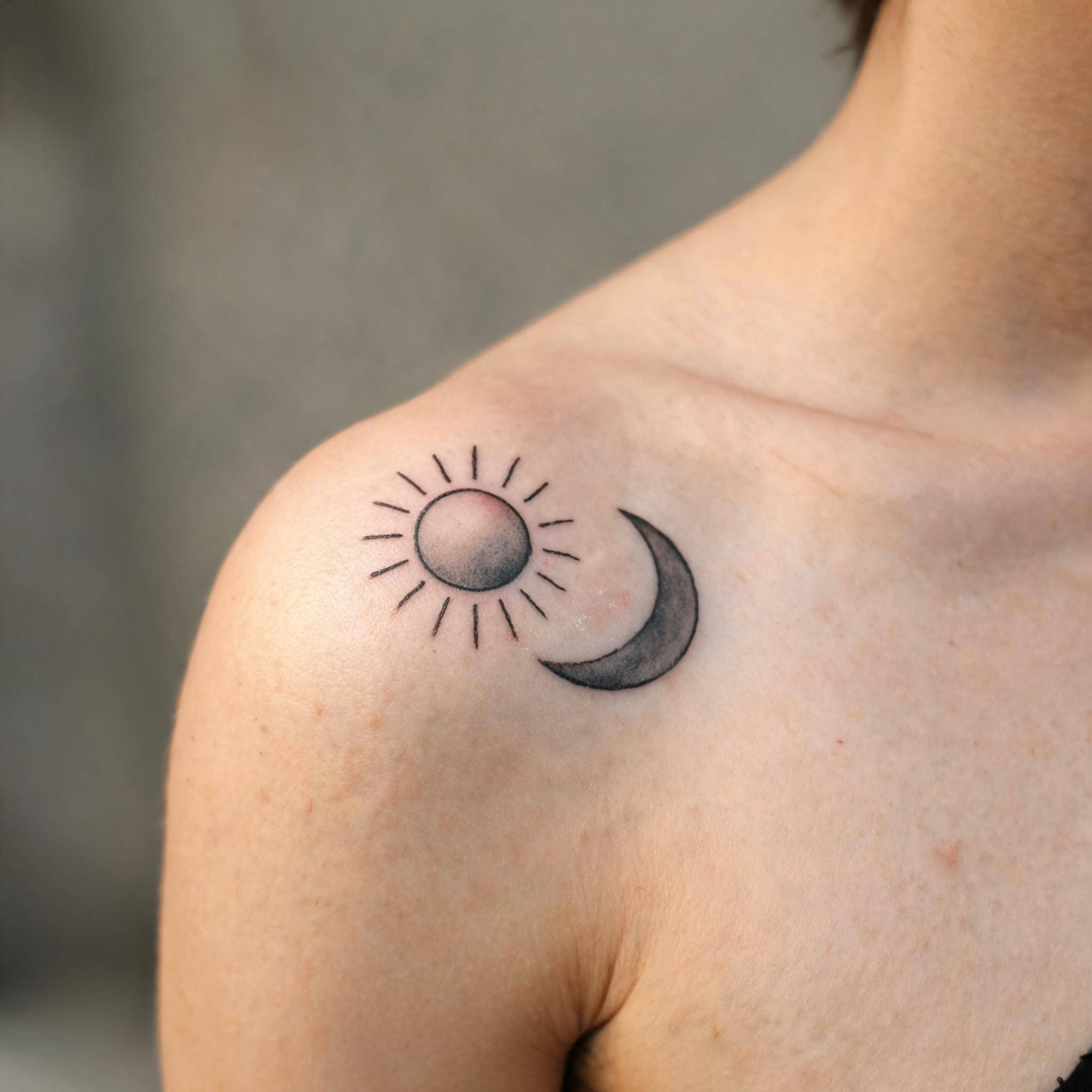 A minimalist sun and moon tattoo image on a shoulder cap, shown in black and grey linework against a plain background.