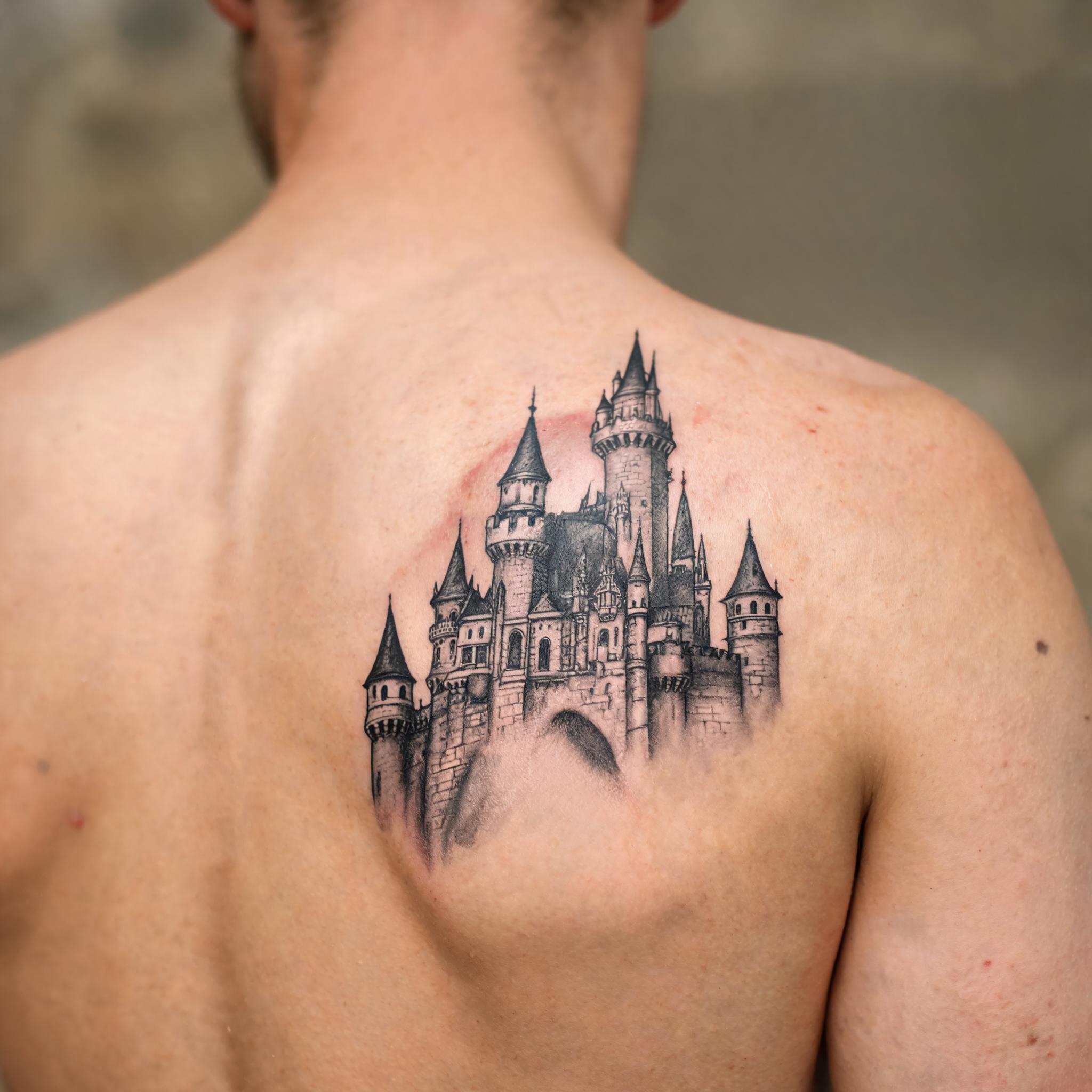 Illustrative black and grey enchanted castle tattoo image on a shoulder blade, shown on realistic skin against a neutral background.