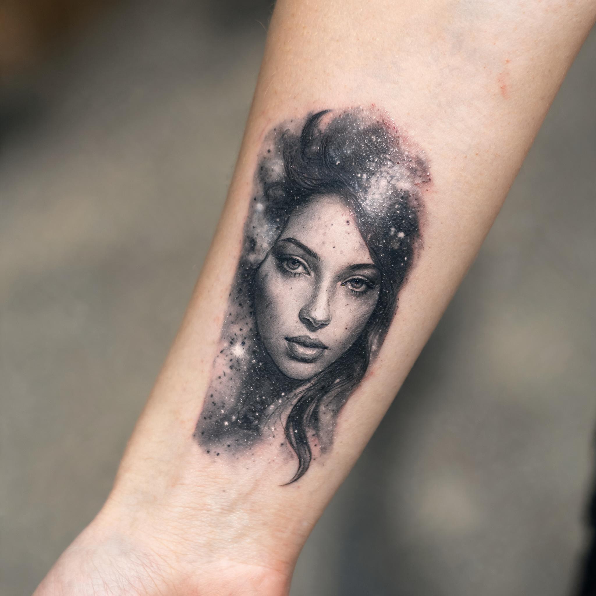 Black and grey realism tattoo image of a cosmic woman face on an outer forearm, shown against a clean neutral background.