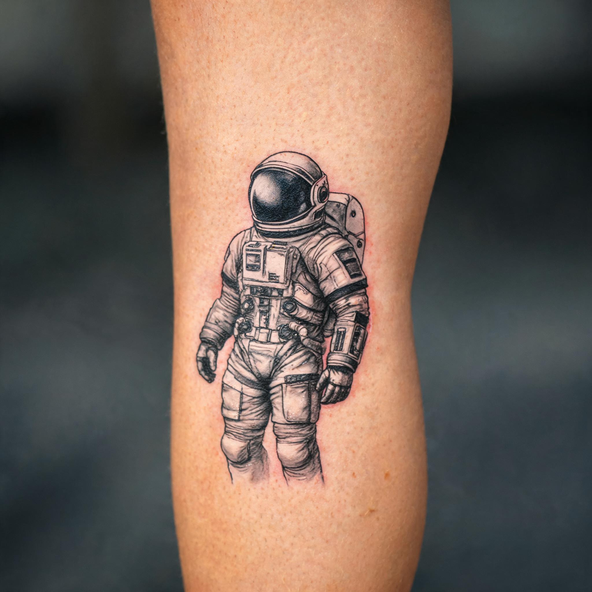 Black and grey realistic astronaut tattoo design shown on a front thigh against a plain background, photographed as a tattoo image.
